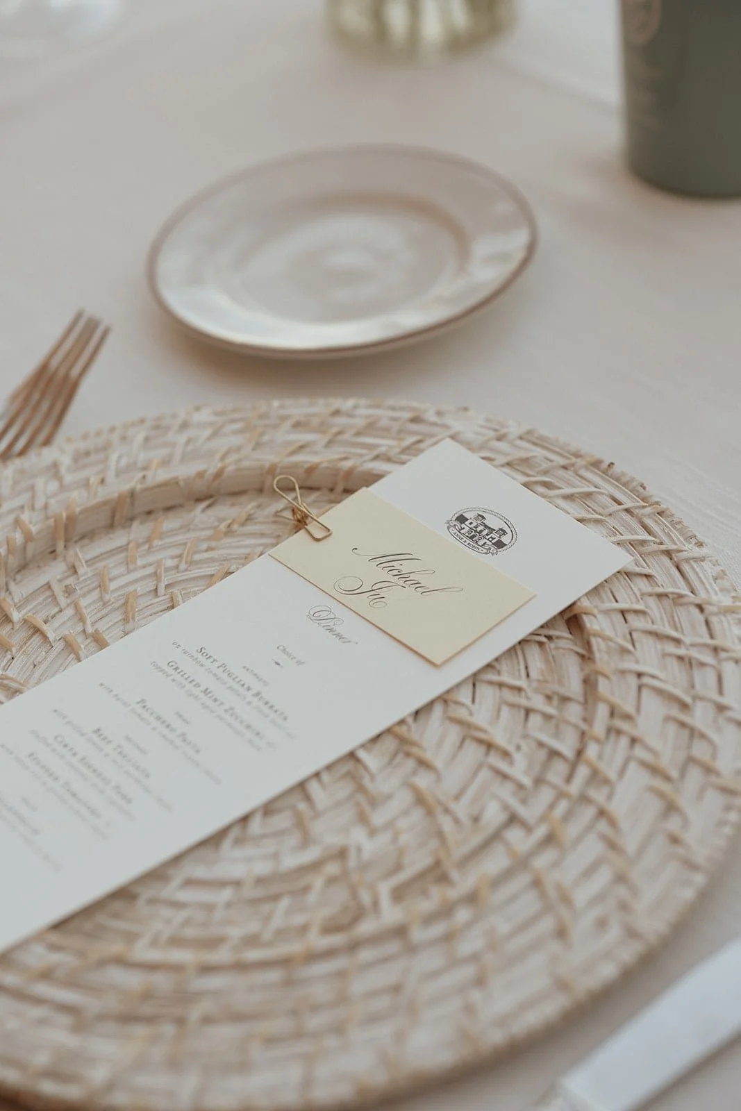 A detailed top-down shot of a place setting featuring a woven rattan charger plate, a custom menu with a gold paperclip name tag for "Michael Jr.," and a small bread plate.