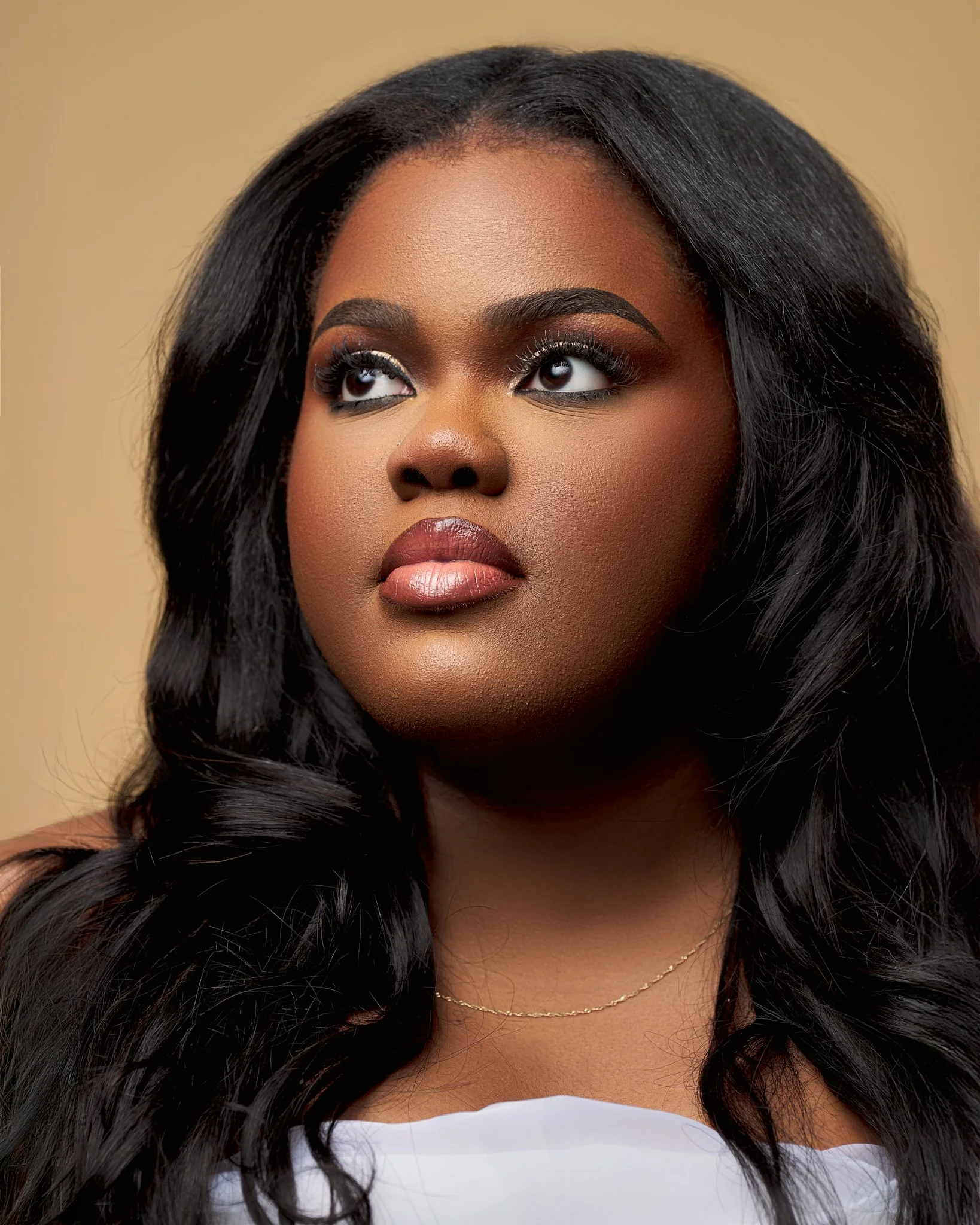 Close-up of a woman with dark hair and makeup looking up, wearing a white top and a gold necklace against a beige background.