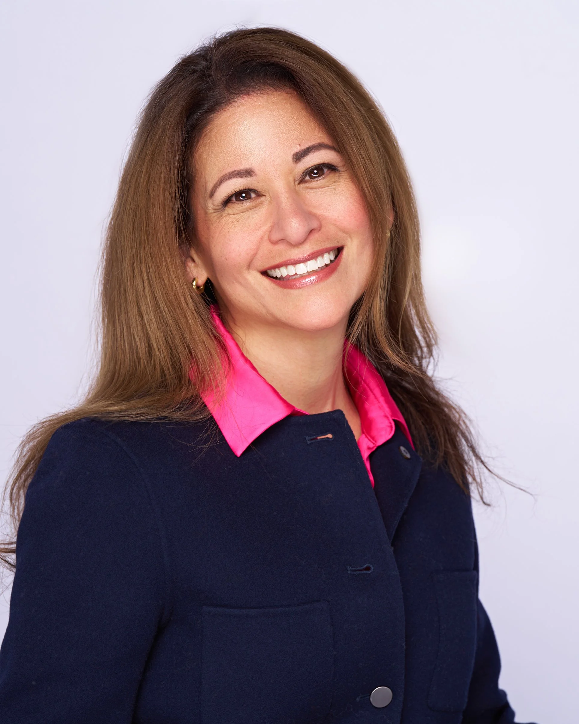 Close-up of a woman with long brown hair smiling, wearing a pink collared shirt and a dark navy blue jacket, against a plain white background.