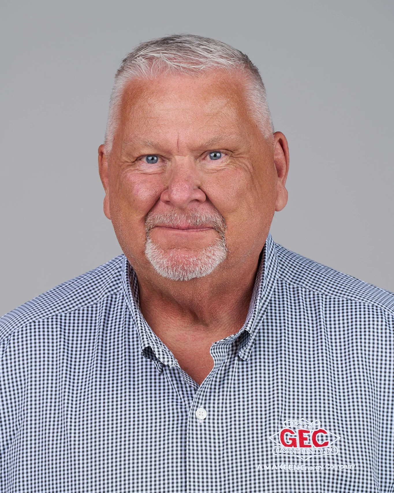 Portrait of an older man with short gray hair, a full gray beard, wearing a checkered shirt, standing against a plain gray background.
