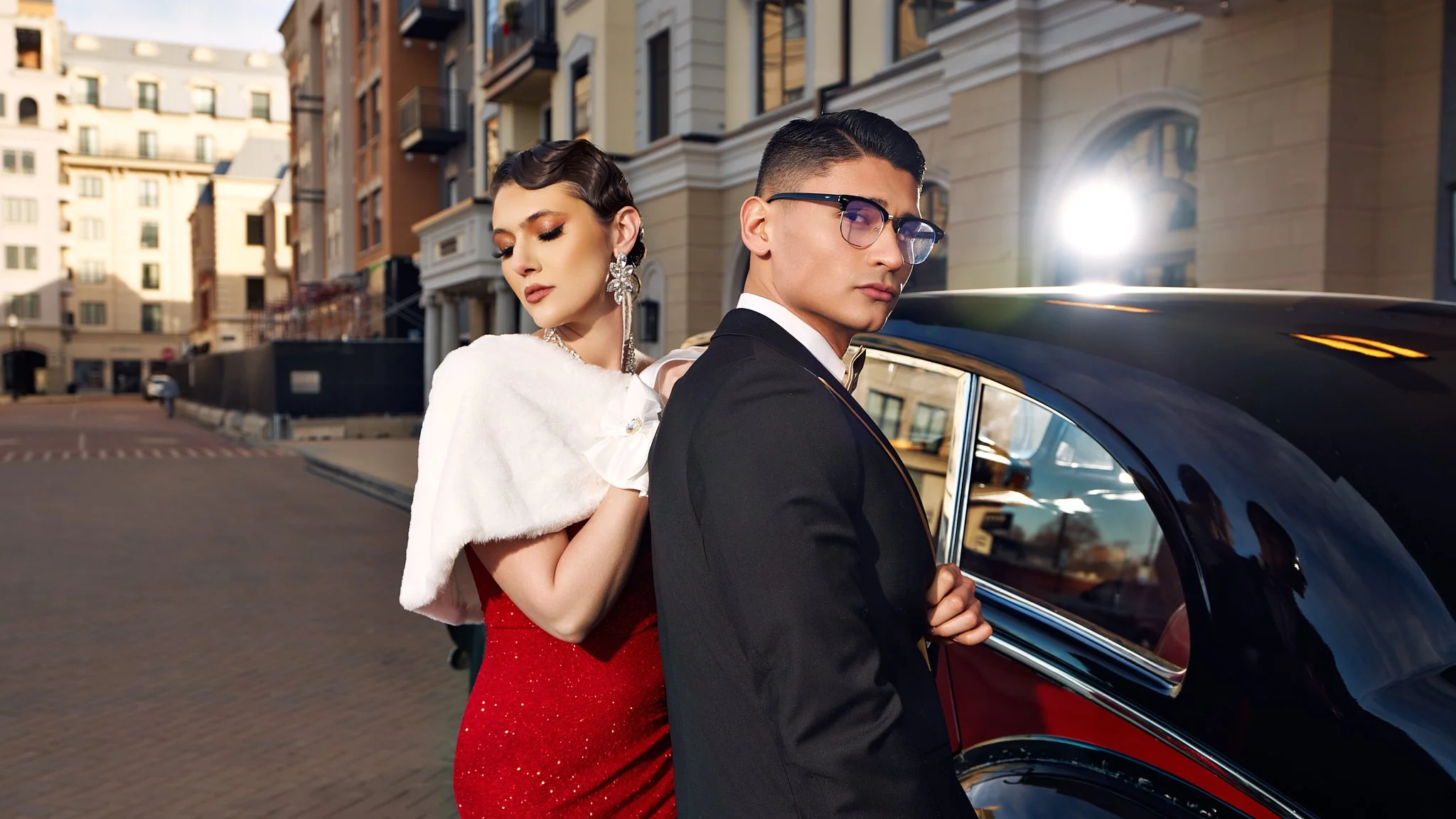 A stylish couple dressed elegantly, standing next to a black luxury car on a city street with buildings and sunlight in the background.