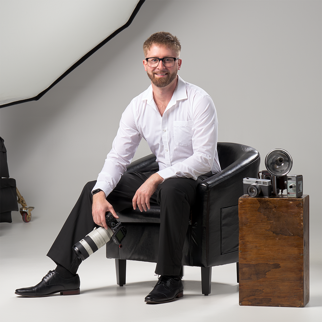 Man sitting on a black leather armchair holding a camera with a long lens, surrounded by vintage cameras and photography equipment in a studio setting.