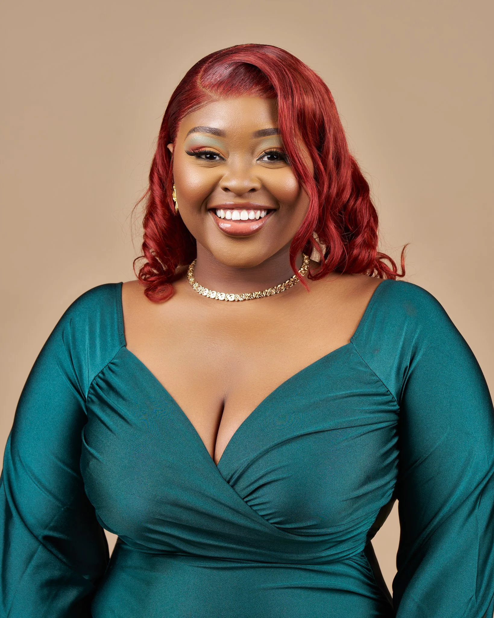 Close-up of an African American woman with red hair styled in loose curls, wearing a teal dress with a deep neckline, gold jewelry, and makeup with gold eyeshadow and a warm lipstick, smiling against a neutral beige background.