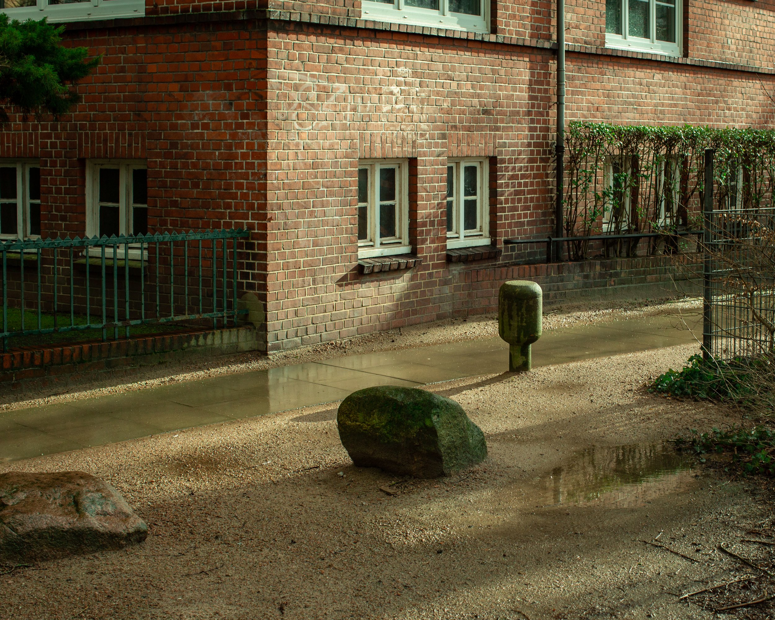 Blick auf eine Ecke eines roten Backsteinhauses mit mehreren kleinen weißen Fenster, umgeben von einem Garten mit Bäumen, Büschen und einer Mauer. Im Vordergrund sind Steine, eine Pfütze und ein Poller auf dem Gehweg sichtbar.