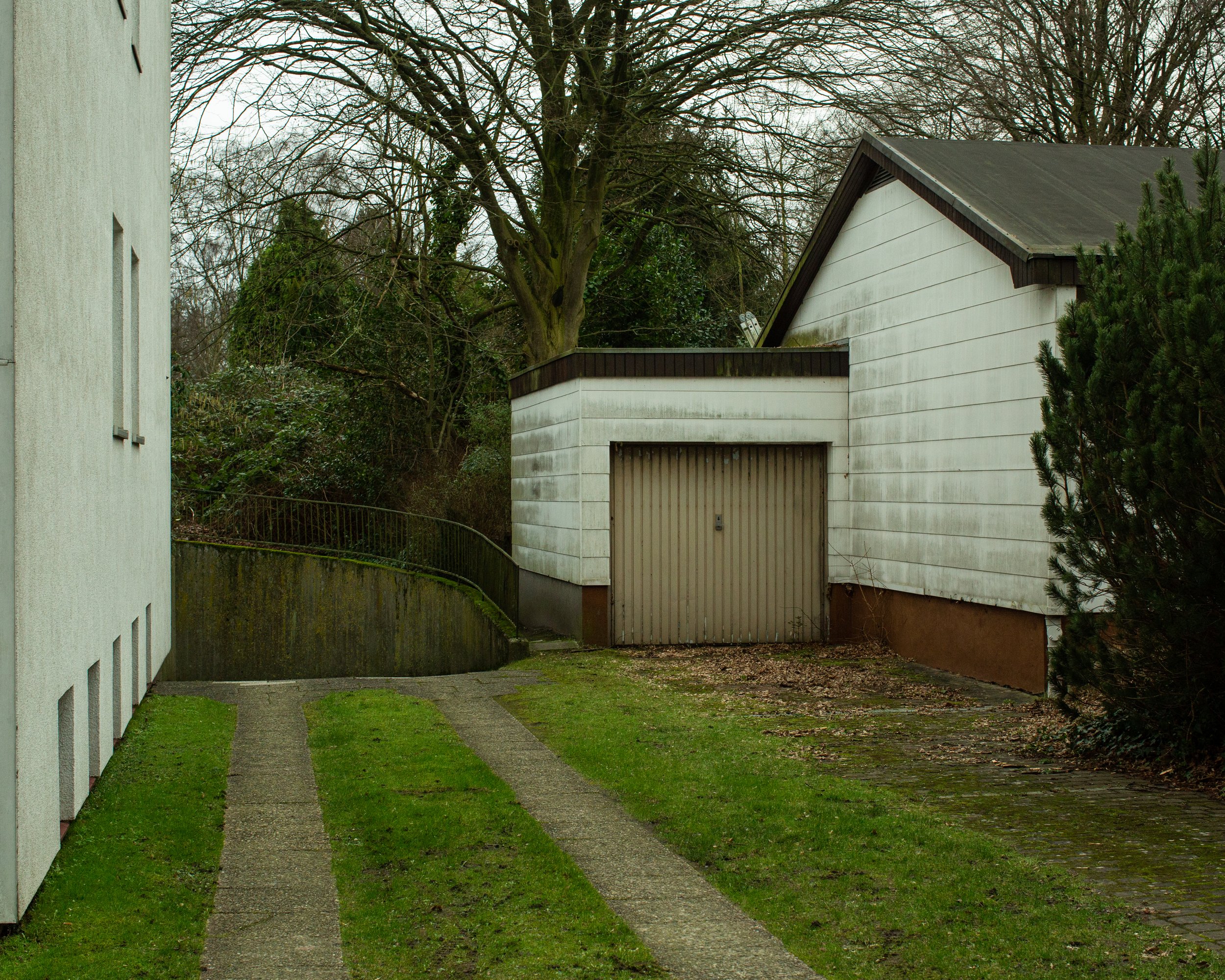 Gebäudeflur, ein weißer Anbau mit Garage und eine gepflegte Rasenfläche mit zwei parallelen Wege. Im Hintergrund Bäume ohne Blätter und ein bewölkter Himmel.