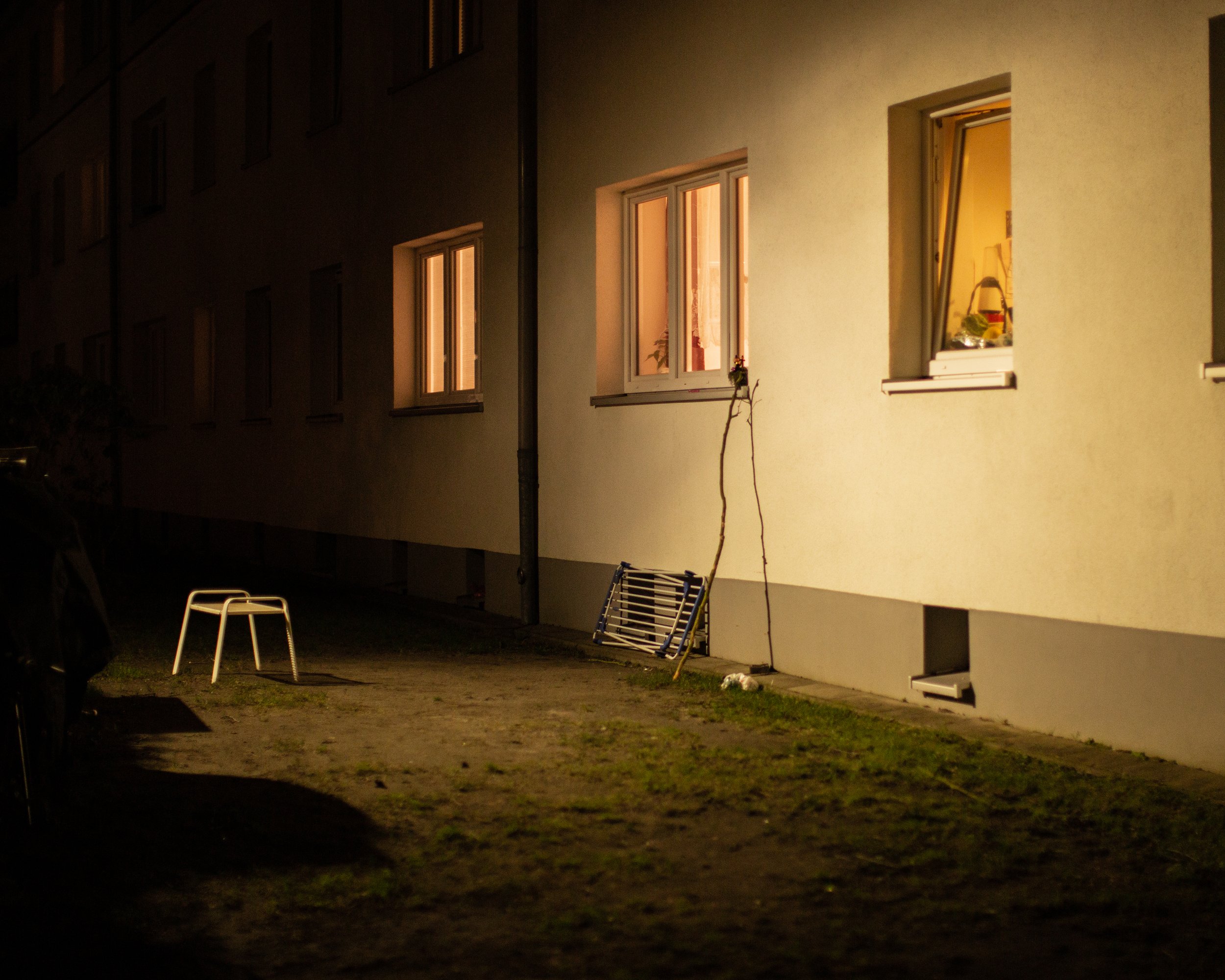 Neontale Seite eines Hauses in der Nacht mit beleuchteten Fenstern, einer weißen Stuhl, einem zusammengelegten Leiter und anderen Gegenständen vor der Wand.