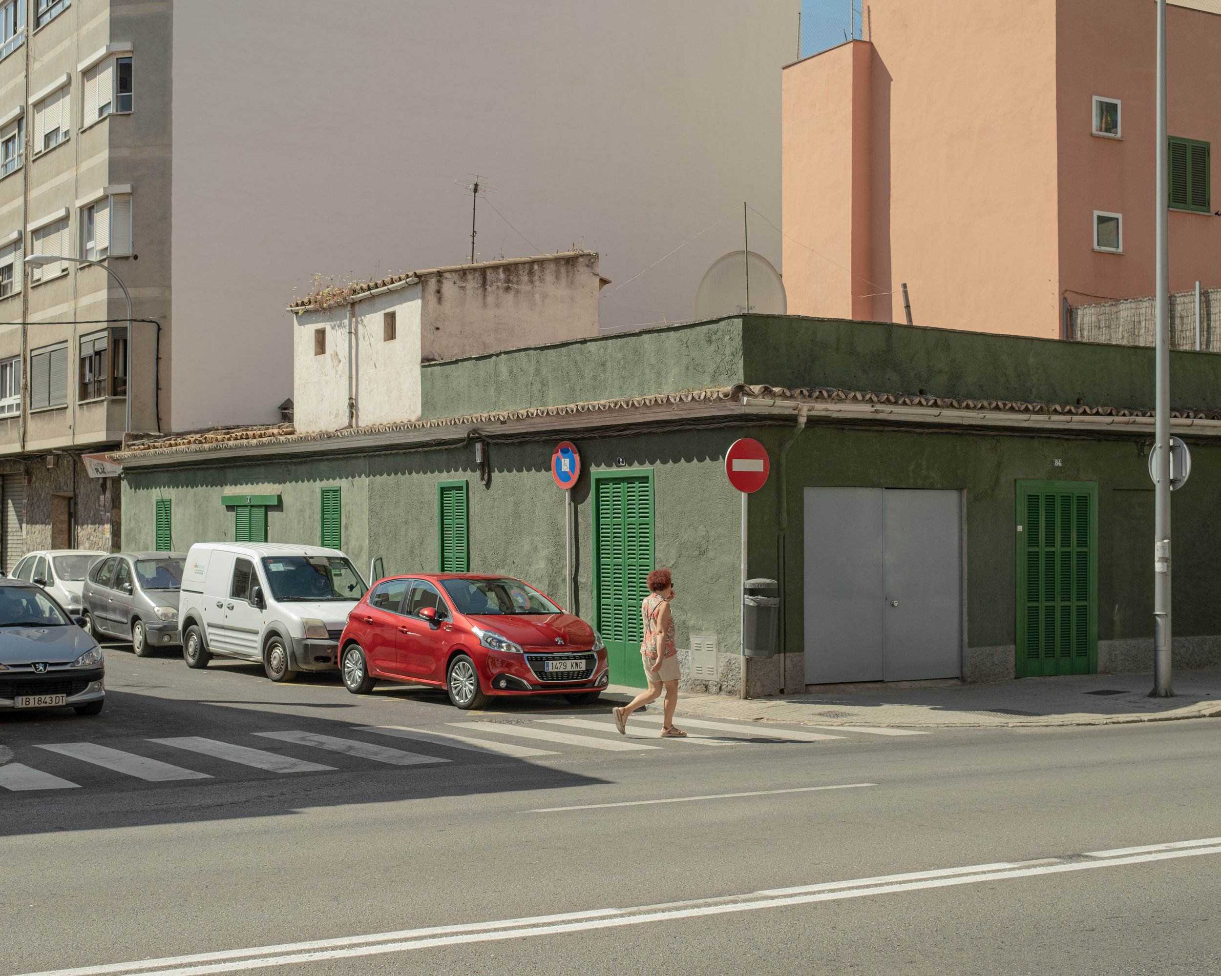 Straße mit parkenden Fahrzeugen, Fußgängerin, grünes Gebäude mit grünen Fensterläden und zwei Straßenschilder, auf einem Gebäude im Hintergrund sind mehrfarbige Wohnhäuser zu sehen.