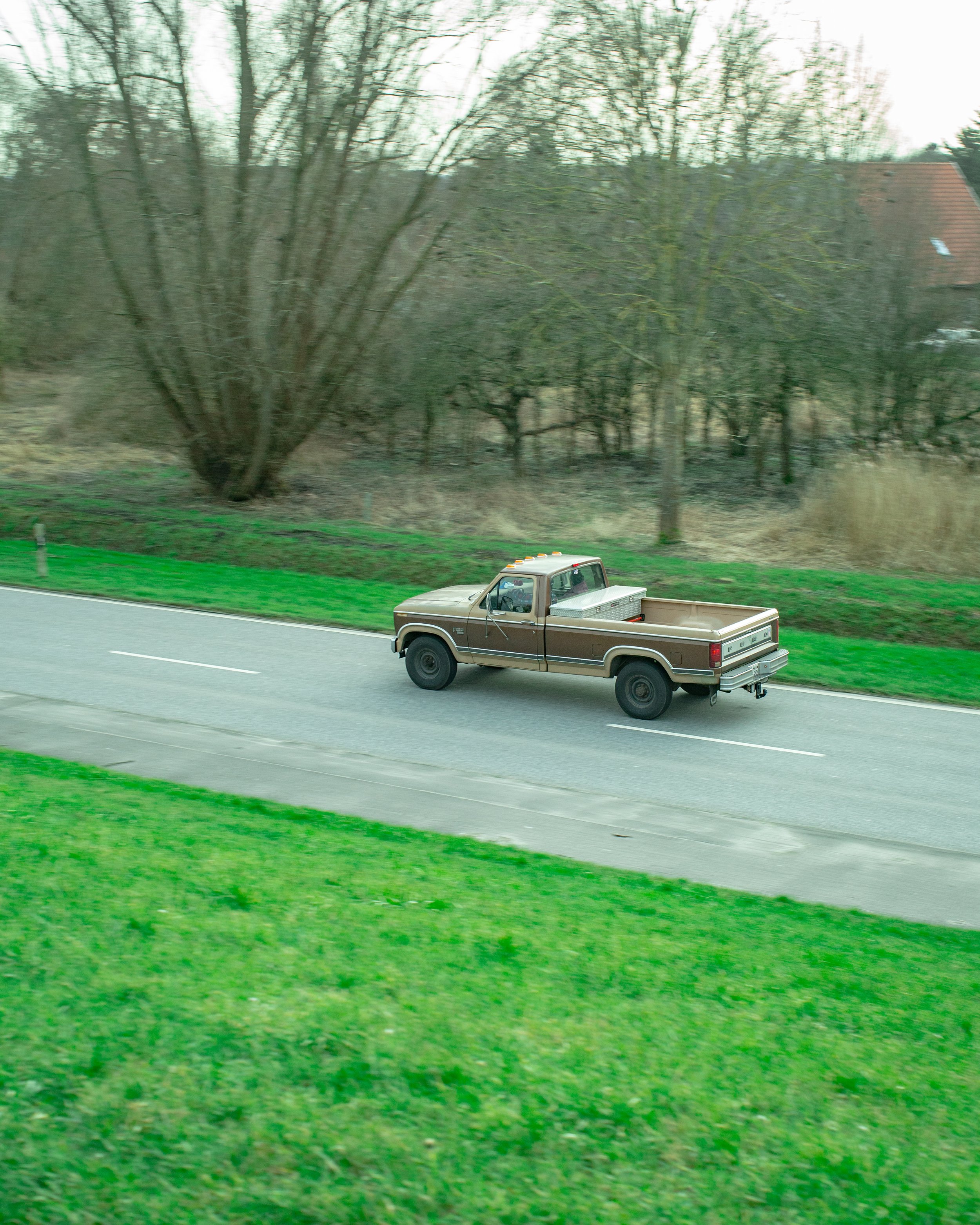 Ein grauer Pickup-Truck fährt auf einer zweispurigen Straße, umgeben von grünem Gras und Bäumen im Hintergrund, bei bewölktem Himmel.
