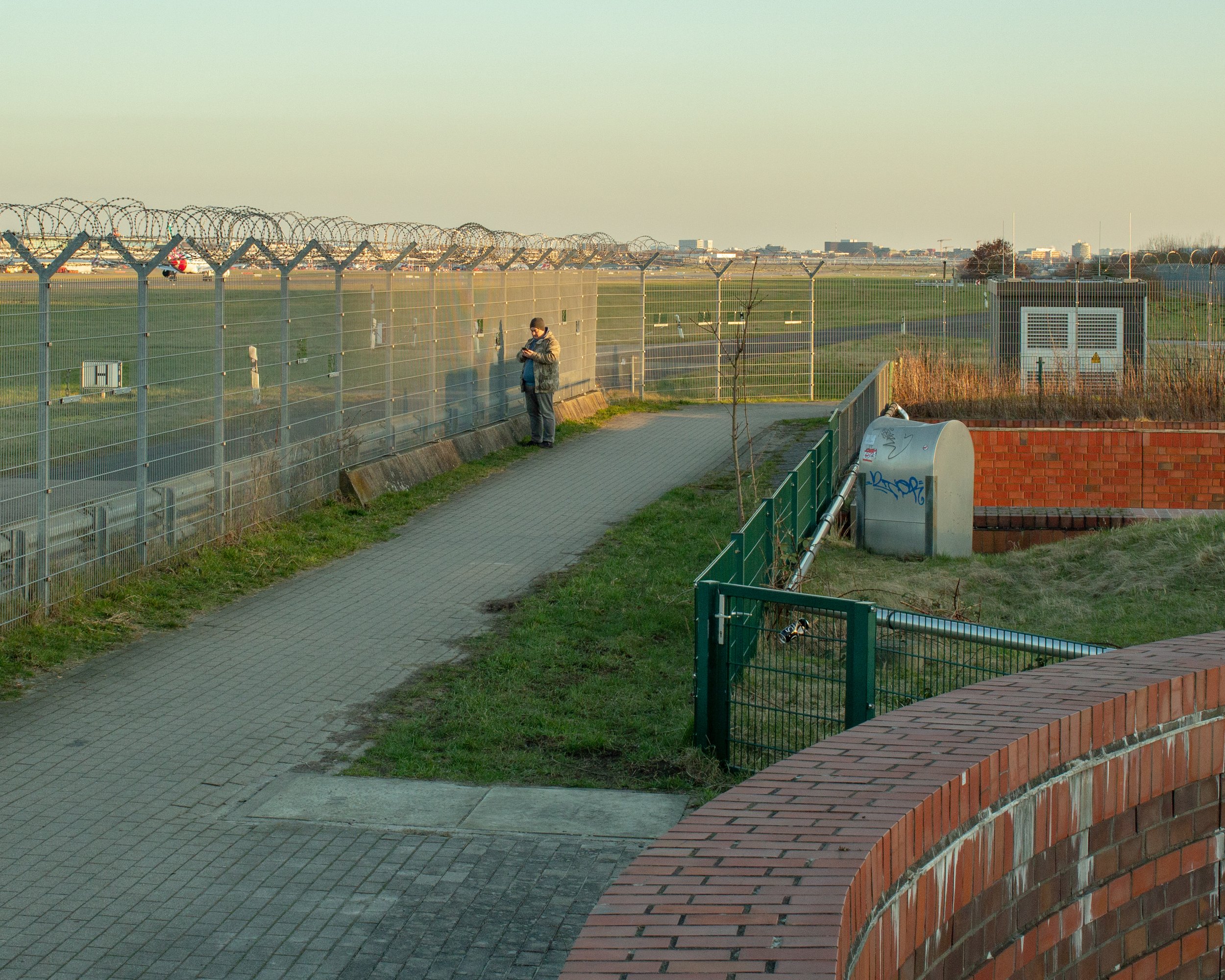 Ein Mann steht an einer Flughafenzäunung und schaut auf sein Handy, im Hintergrund sind Flugzeuge auf dem Rollfeld sichtbar.