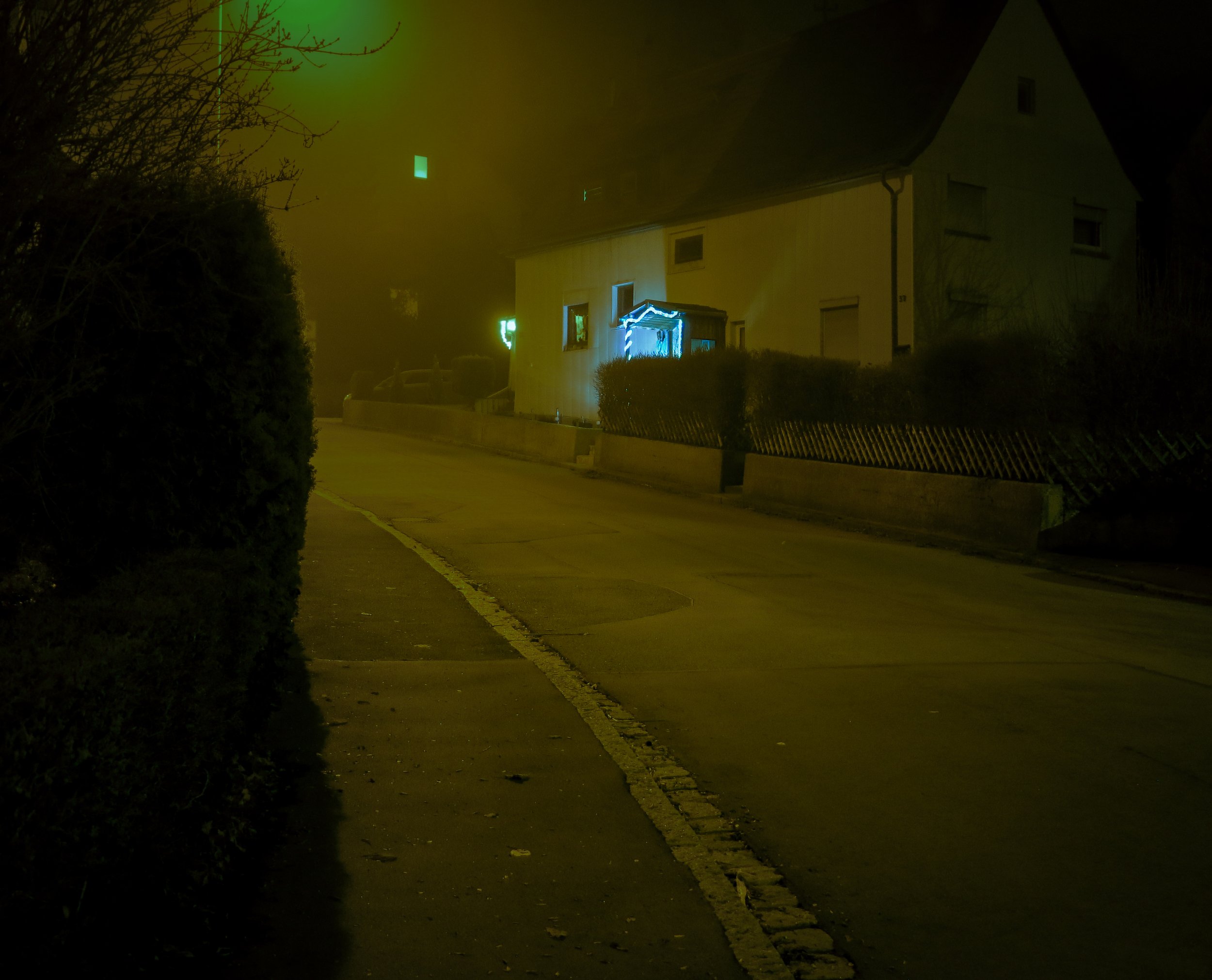 Dunkle Straße bei Nacht mit Haus, das mit blauer Lichterdeko geschmückt ist, und Nebel in der Luft.
