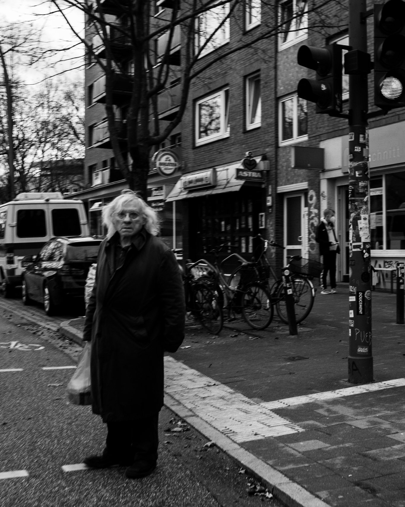 Ein älterer Mann mit langen Haaren und Brille steht an einer Straßenkreuzung in einer Stadt, umgeben von geparkten Fahrrädern, Autos und Gebäuden. Es ist schwarz-weiß fotografiert.