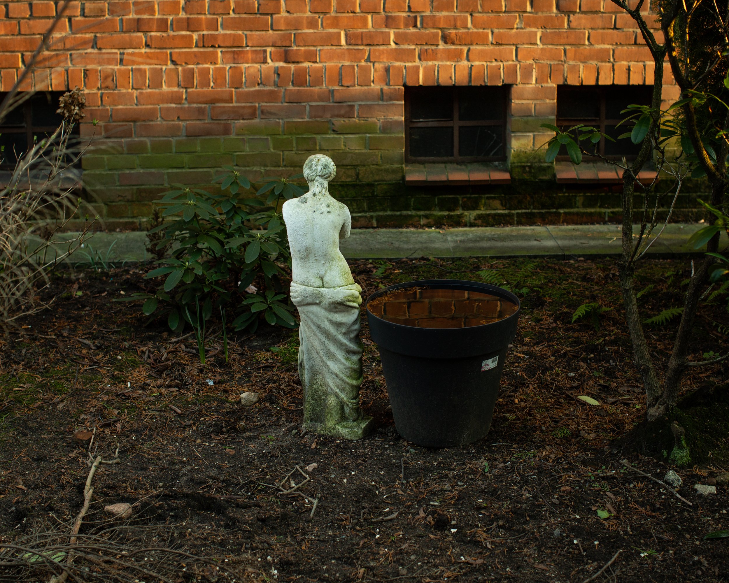 Garten mit einer kleinen Skulptur einer nackten Frau und einem schwarzen Blumentopf, der mit Wasser gefüllt ist, vor einem Backsteinhaus