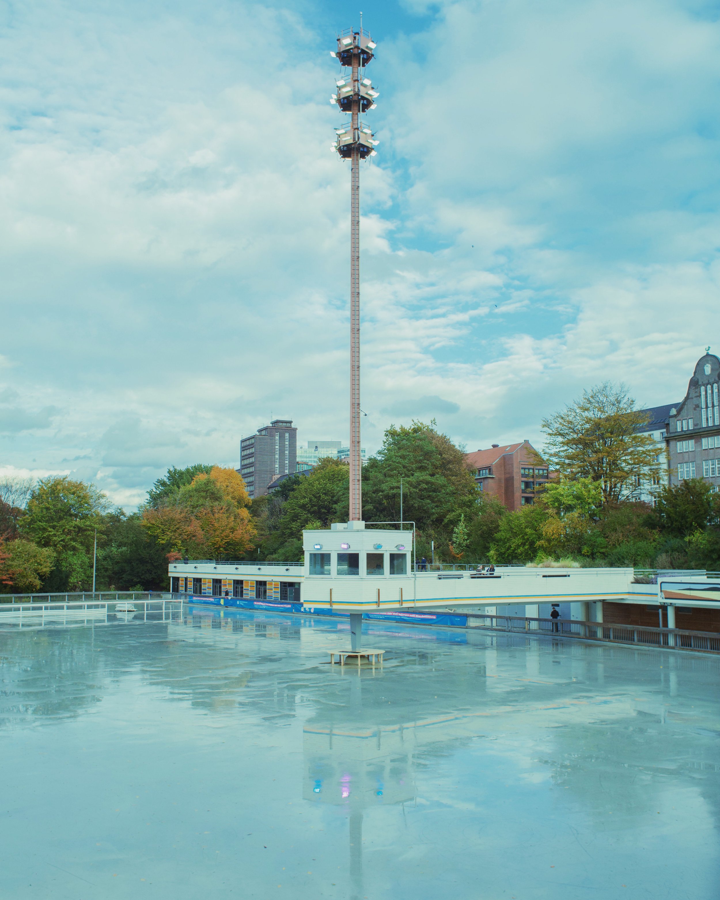 Eislauffläche mit Wasser, eine kleine Gebäude mit einem hohen Antennenturm, dahinter Bäume und Hochhäuser bei bewölktem Himmel.