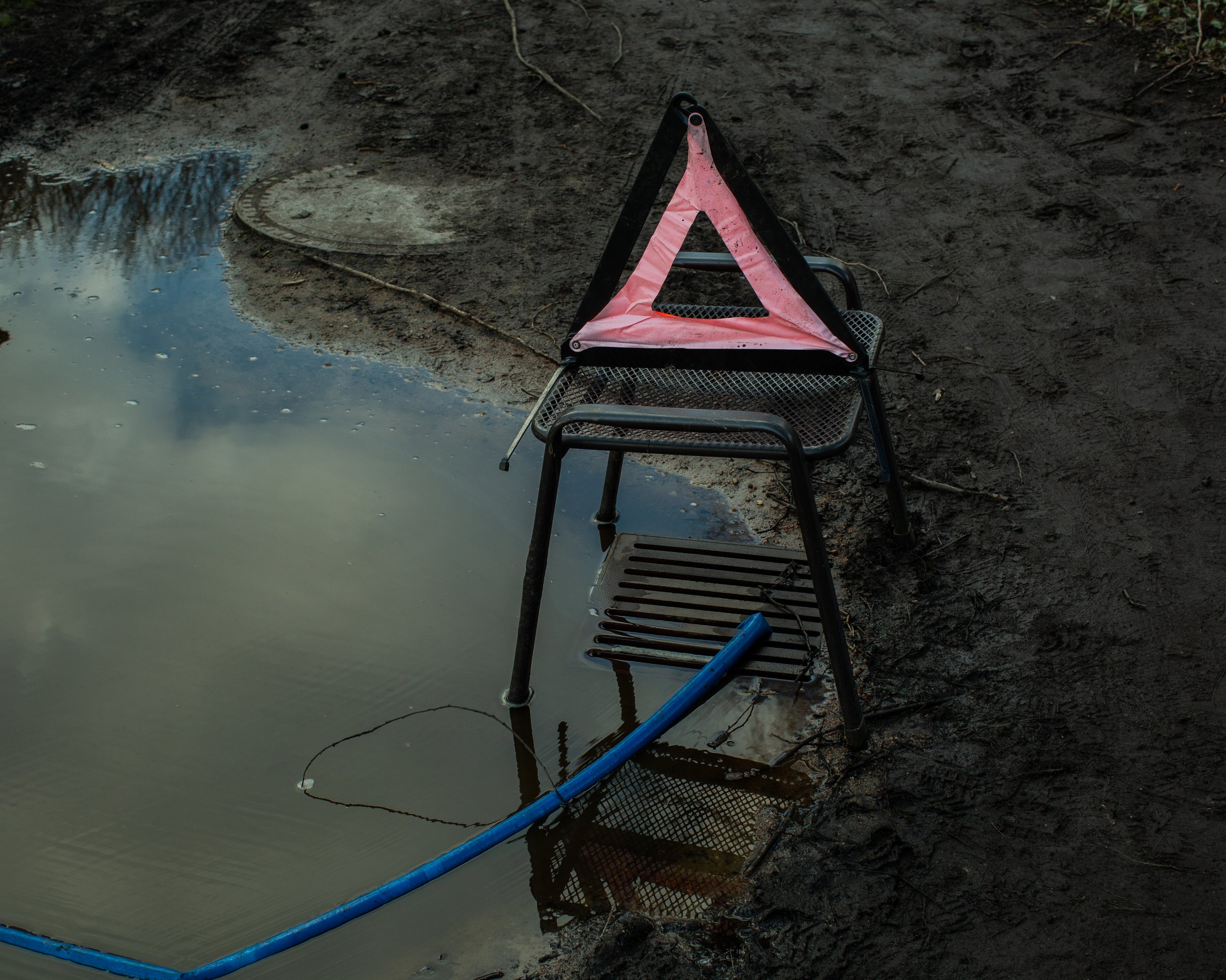 Ein umgestürzter Stuhl mit einem Sicherheitsdreieck auf der Sitzfläche, liegt am Ufer eines Teiches, das Wasser ist ruhig und spiegelt den Himmel wider.