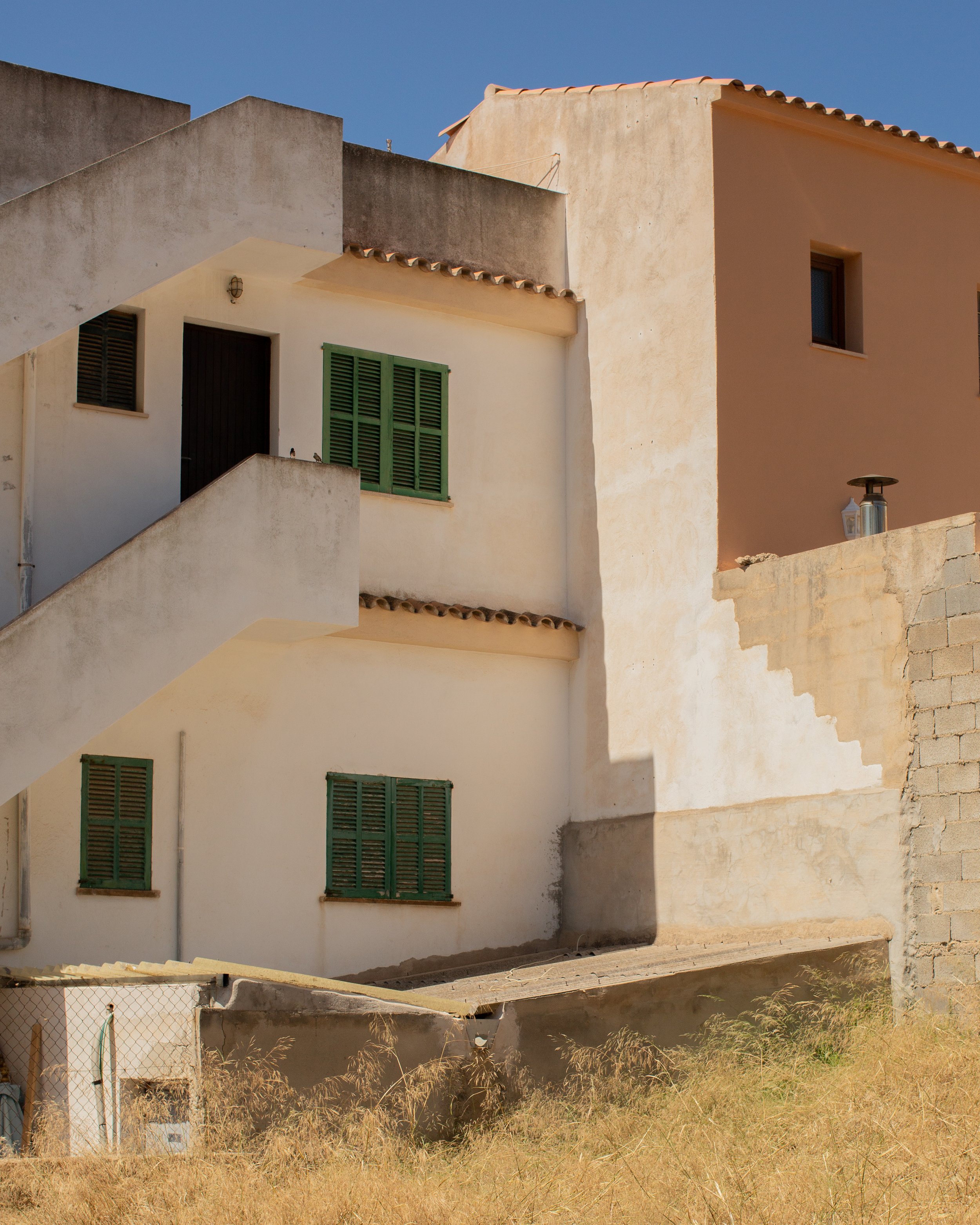 Mehrstöckiges Gebäude mit weißen Wänden, grünen Fensterläden, Außentreppen und einem ummauerten Bereich mit Gras davor, bei Sonnenlicht, blauer Himmel.