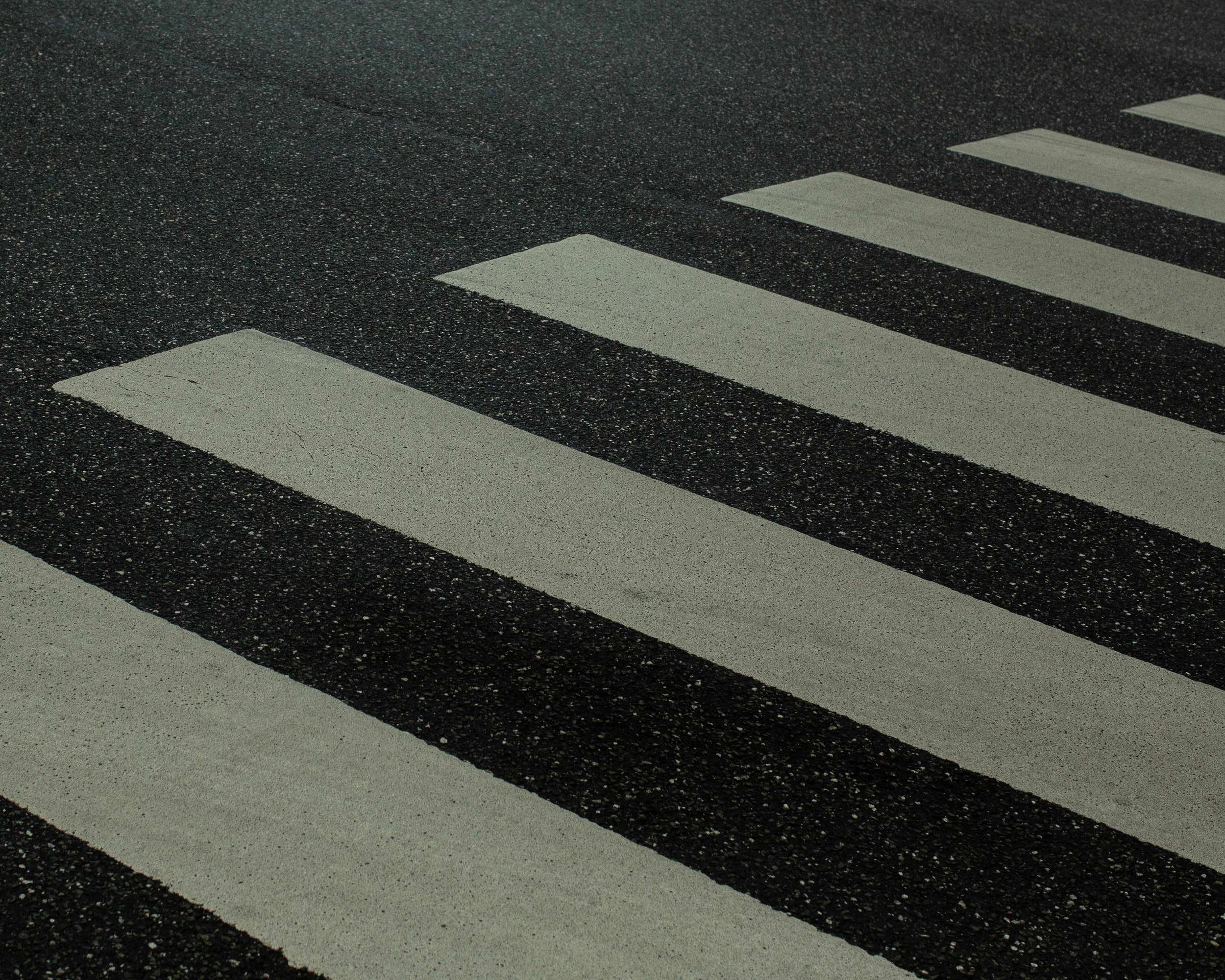 Nahaufnahme eines Zebrastreifen auf einer asphaltierter Straße, bestehend aus weißen Streifen auf dunklem Asphalt.