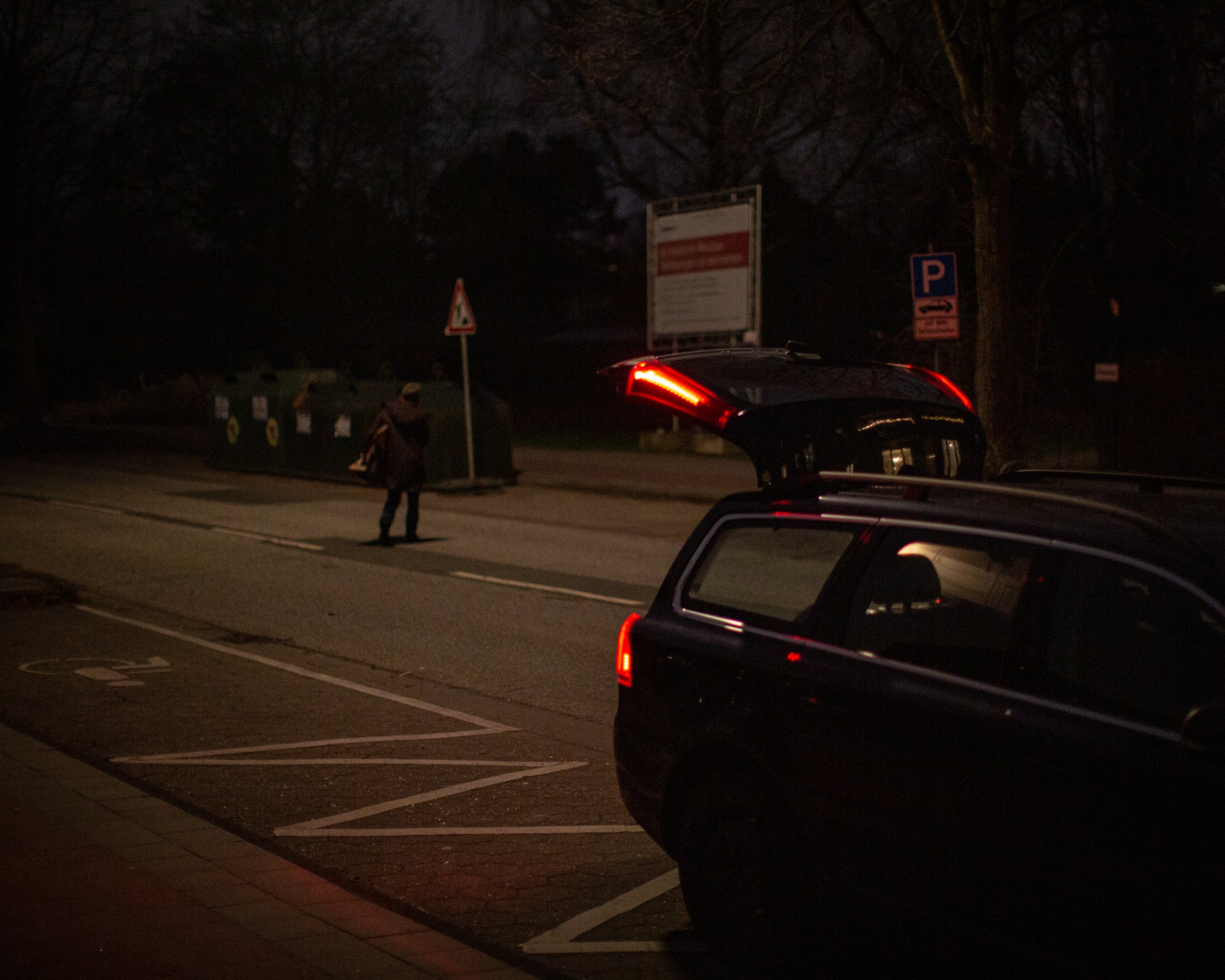 Ein beleuchteter Parkplatz bei Nacht mit einem schwarzen Fahrzeug im Vordergrund, das offene Kofferraum hat. Im Hintergrund steht eine Person an der Straße, neben Verkehrsschildern und einem Papierkorb. Es sind Bäume ohne Blätter sichtbar, die den Hi