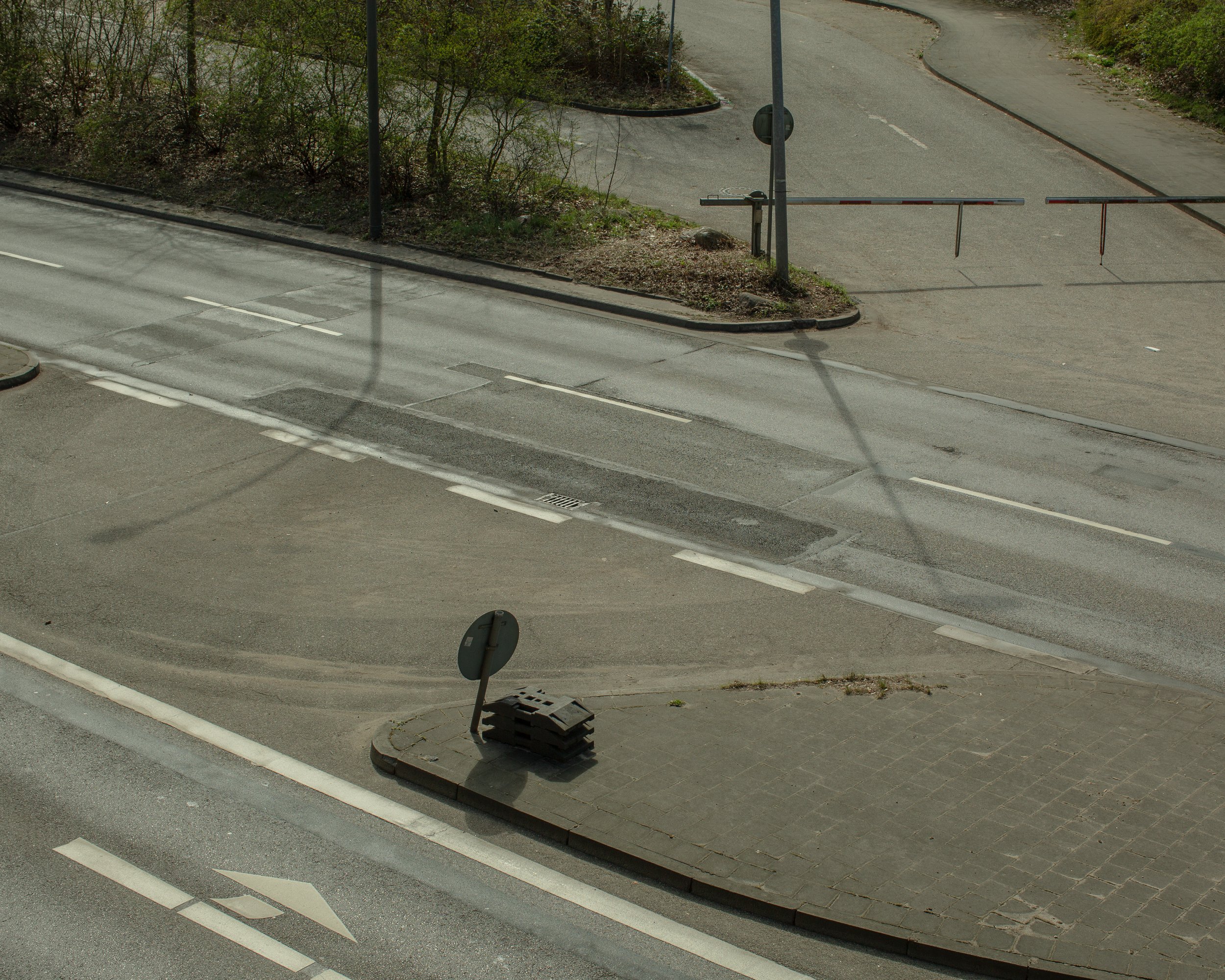 Leerer Straßenabschnitt mit einem Fahrbahnmarkierung, einem kleinen gepflasterten Bereich, einigen Verkehrszeichen und einem Absperrzaun im Hintergrund, Schatten von einem Verkehrszeichen auf der Straße