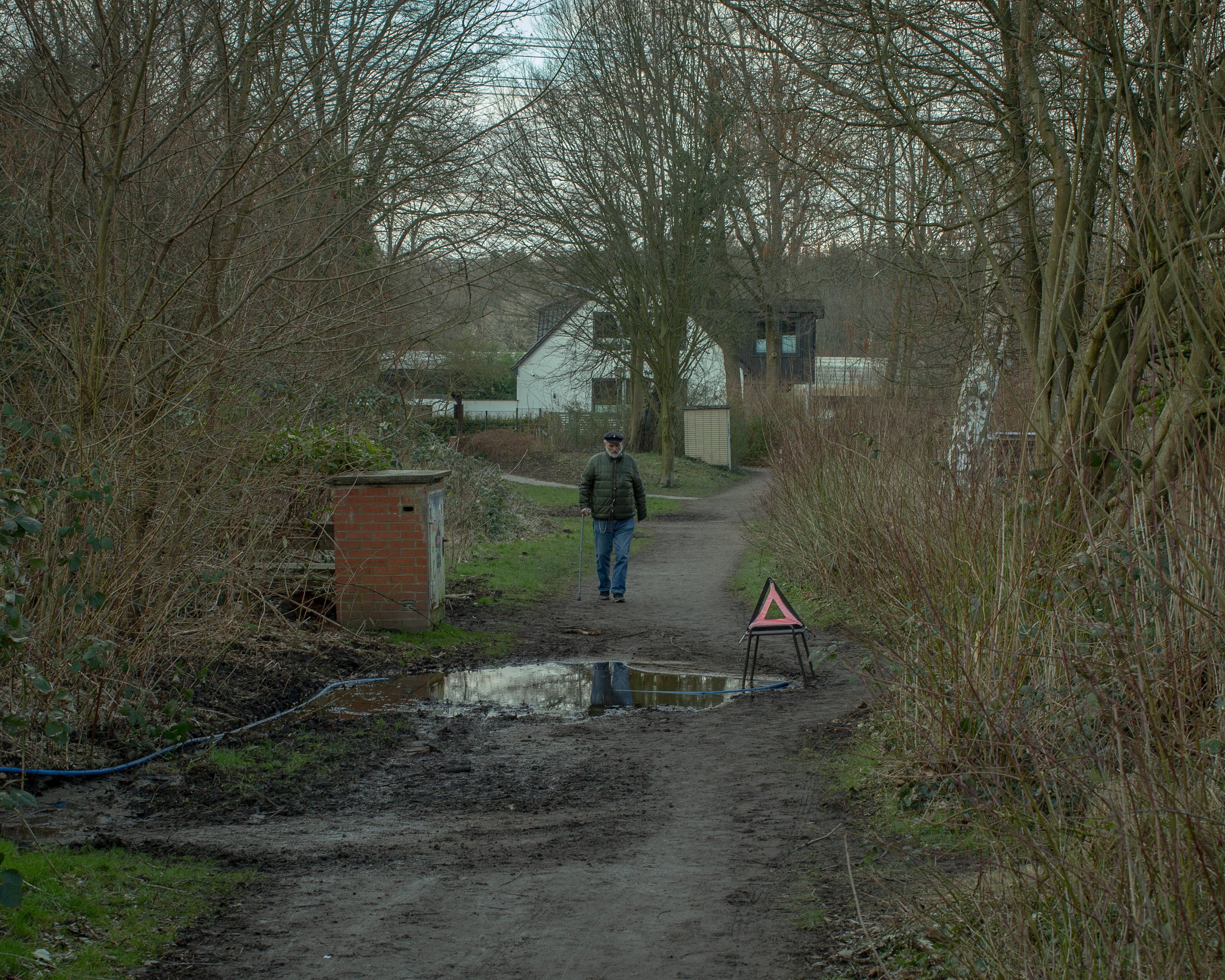 Ein Mann mit einem Spazierstock geht eine schmutzige, unbefestigte Straße entlang. Neben ihm steht ein Warnschild, und ein Pfütze befindet sich auf dem Weg. Umgeben von Büschen und Bäumen, mit Wohnhäusern im Hintergrund, bei diffusem Tageslicht.