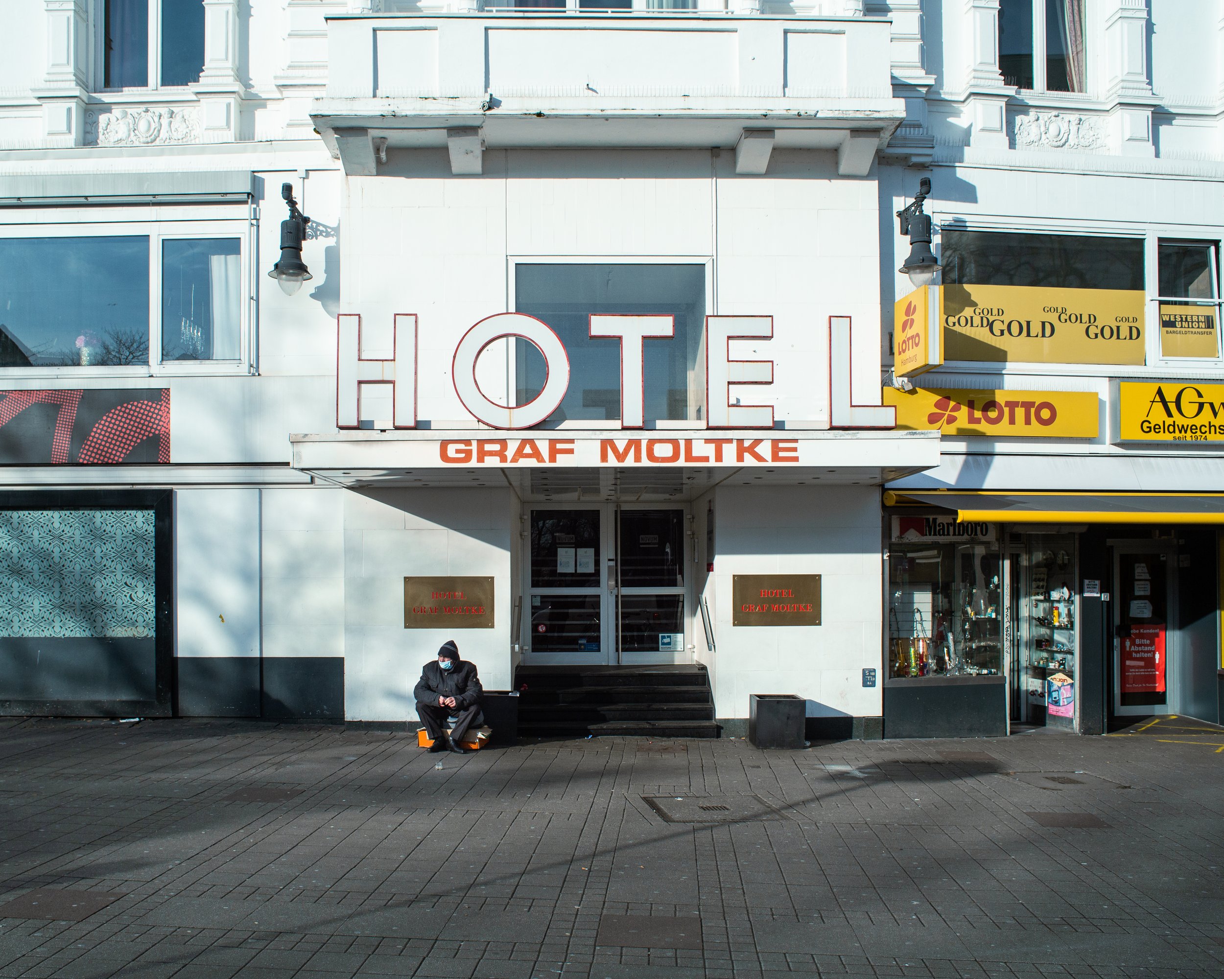 Das Bild zeigt ein Hotelgebäude mit einem großen roten Schriftzug 'HOTEL' und darunter die Bezeichnung 'GRAF MOLKE'. Es gibt eine Person, die auf einer Bank vor dem Hotel sitzt, und Geschäfte auf beiden Seiten der Eingangstür, darunter ein Lotto-Gesc