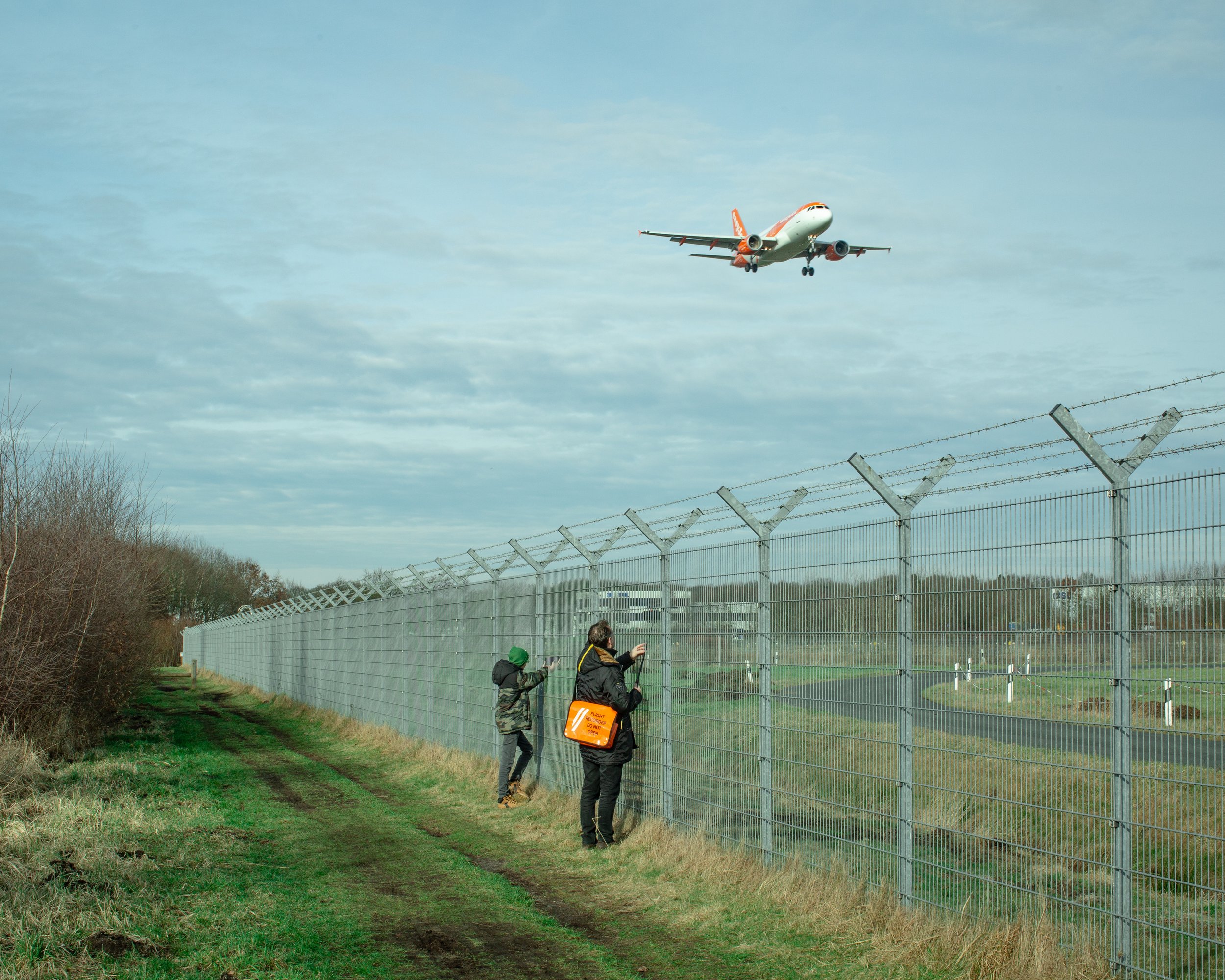 Zwei Personen beobachten ein landendes Flugzeug durch einen Zaun zur Sicherheitskontrolle an einem Flughafen, im Hintergrund ein Himmel mit Wolken.