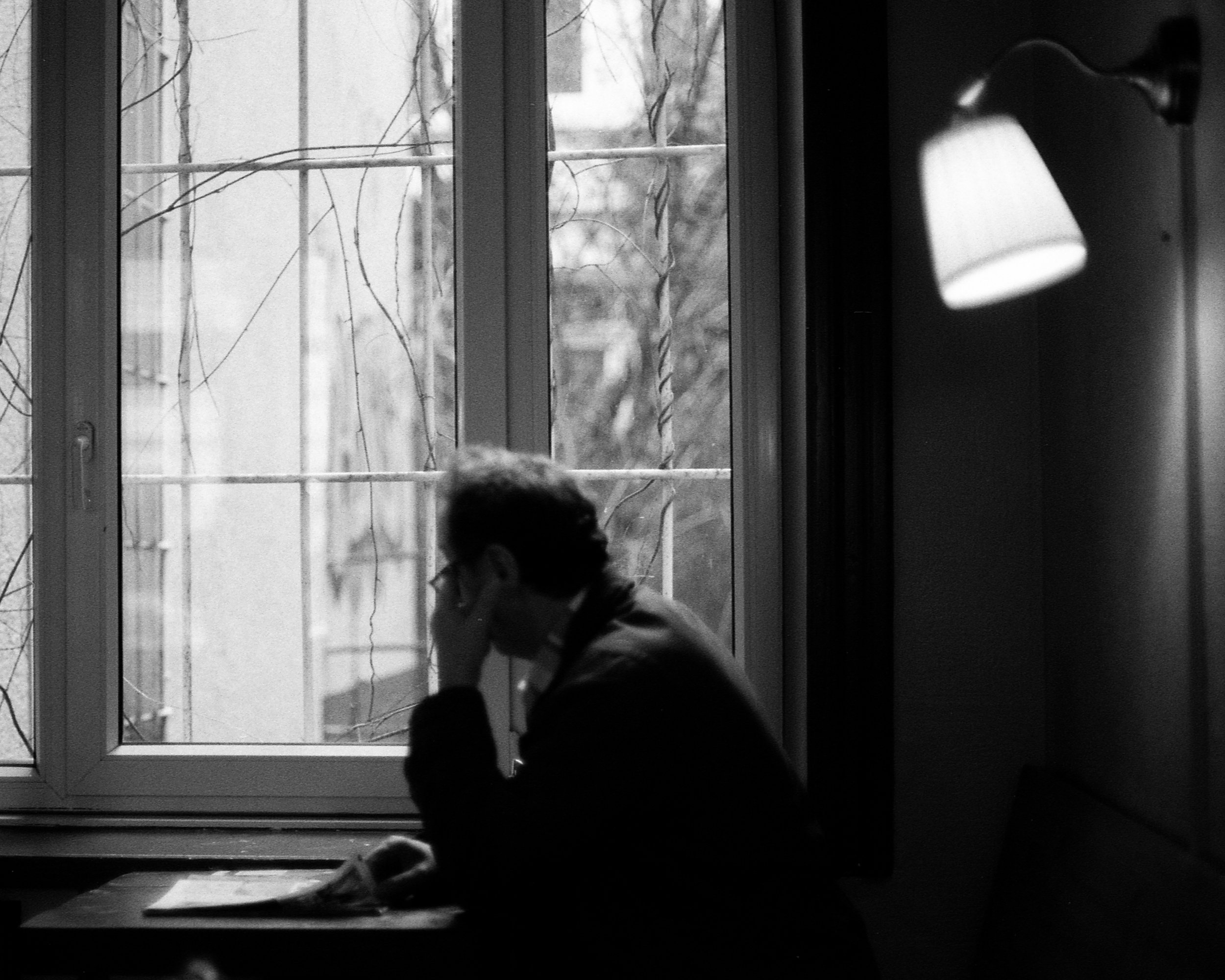 Eine Person sitzt nachdenklich vor einem Fenster im schwarz-weißen Foto, wirkt nachdenklich, mit der Hand am Kopf, im Innenraum mit einer Wandlampe an der Wand.