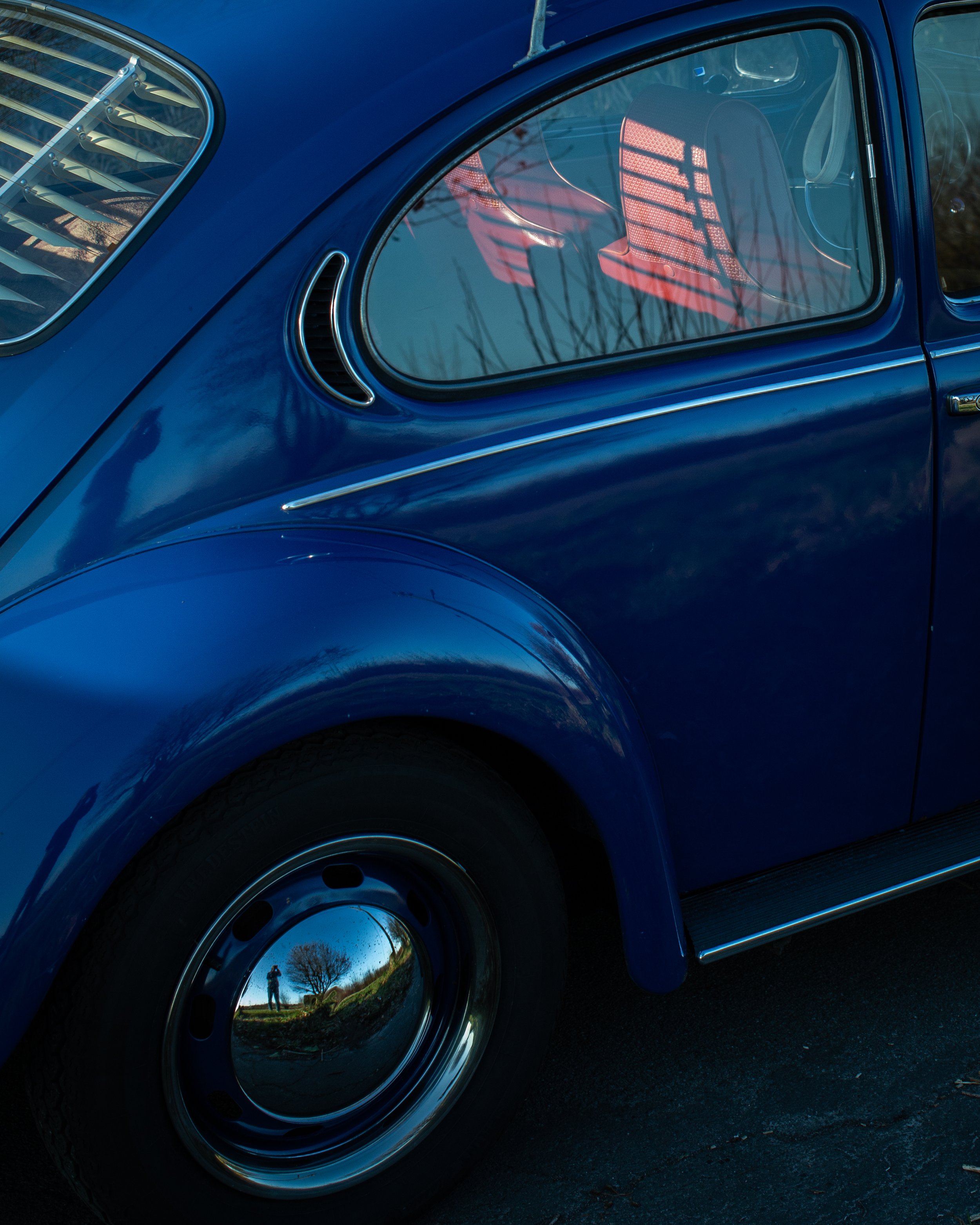 Nahaufnahme eines blauen Vintage Autos mit reflektierendem Rad und Fenstern, Spiegel zeigt die Silhouette eines Baumes und eine Person