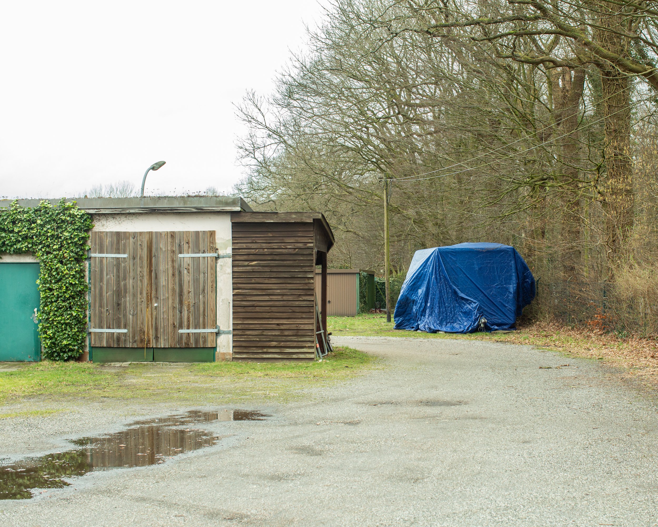 Ein umgebauter Schuppen mit einer dunklen Holzwand und weiteren Gebäuden dahinter, daneben eine mit einem blauen Planen bedeckte offizielle Fahrzeug- oder Geräteabdeckung, umgeben von Bäumen und einer leicht nassen asphaltierten Fläche.