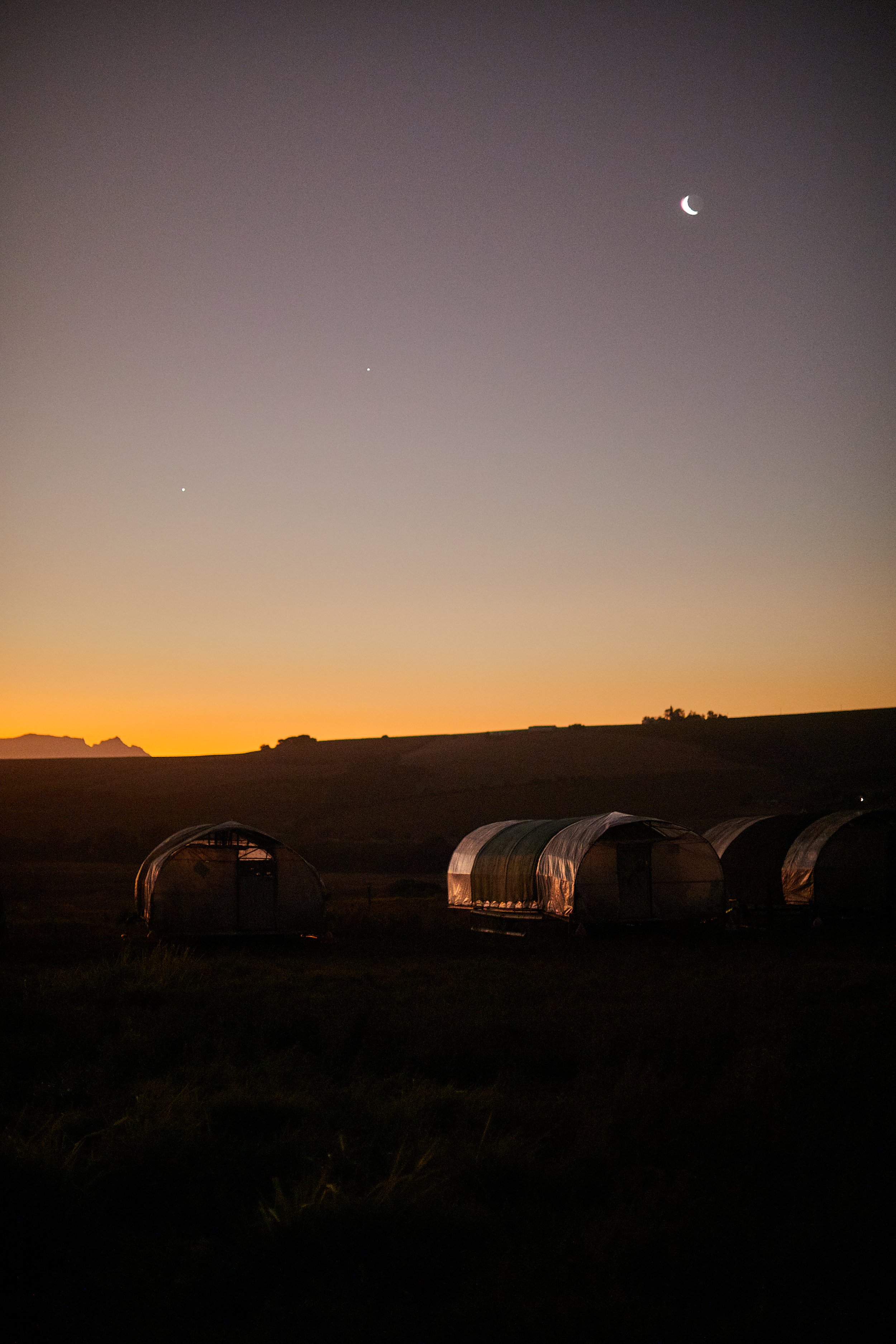 Sternenhimmel bei Sonnenuntergang über Zelten auf einer Wiese, mit sichtbarem Mond und Planeten.