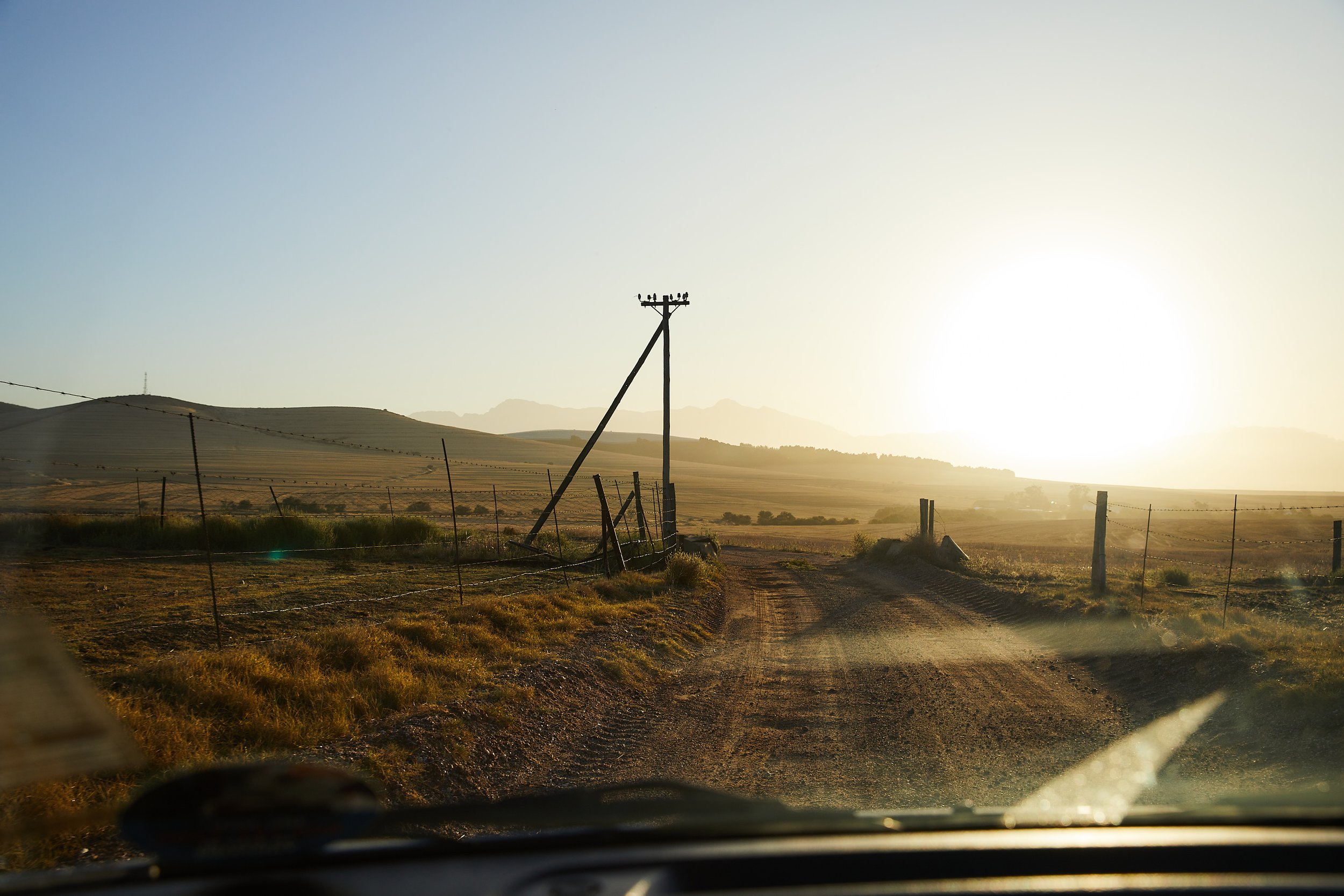 Ländliche unbefestigte Straße bei Sonnenuntergang, umliegende Felder, Holzpfosten, und eine pastoral wirkende Landschaft.