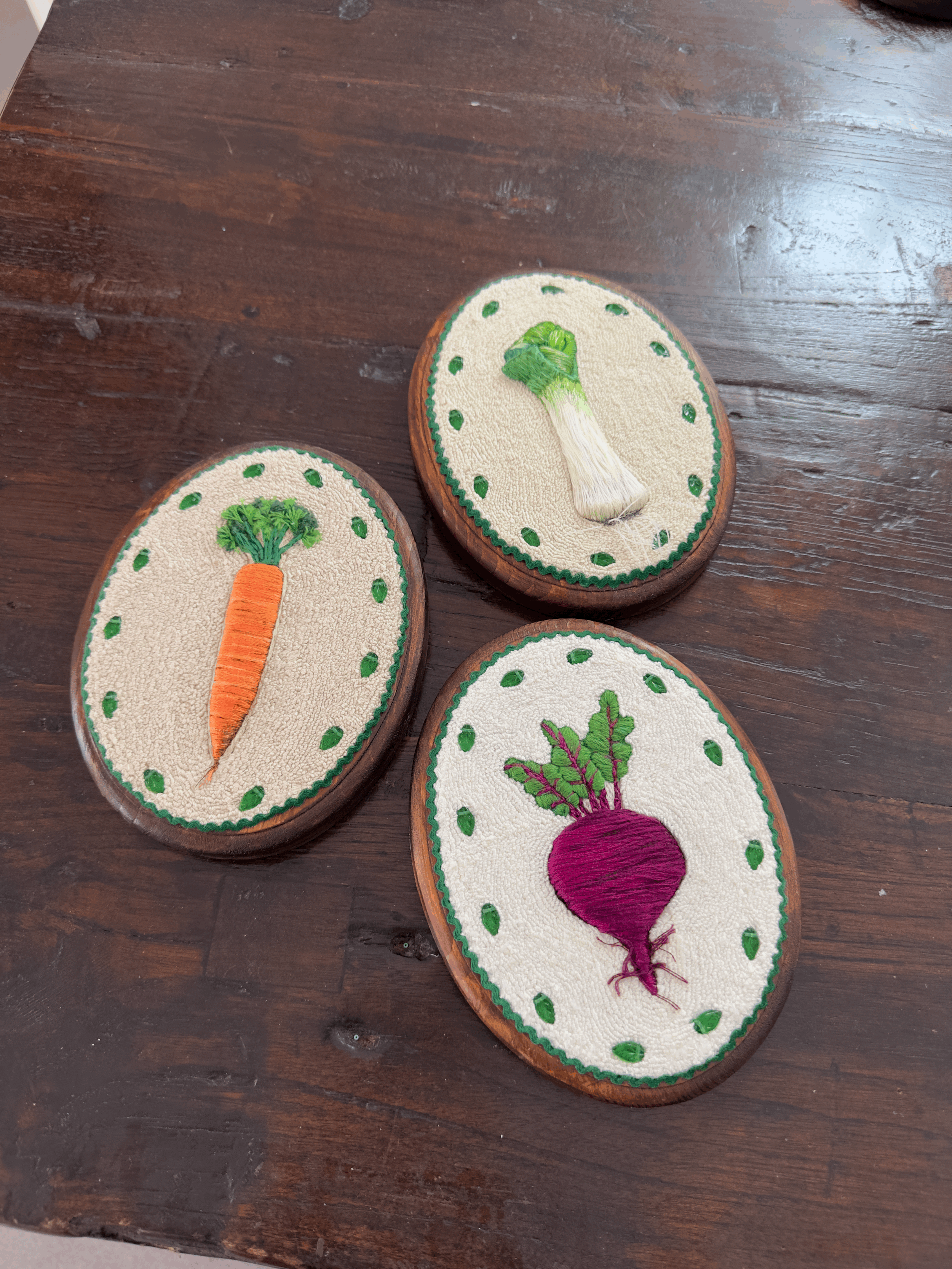 Three embroidered wooden plaques with vegetable designs: a carrot, a beet, and a broccoli, placed on a wooden surface.