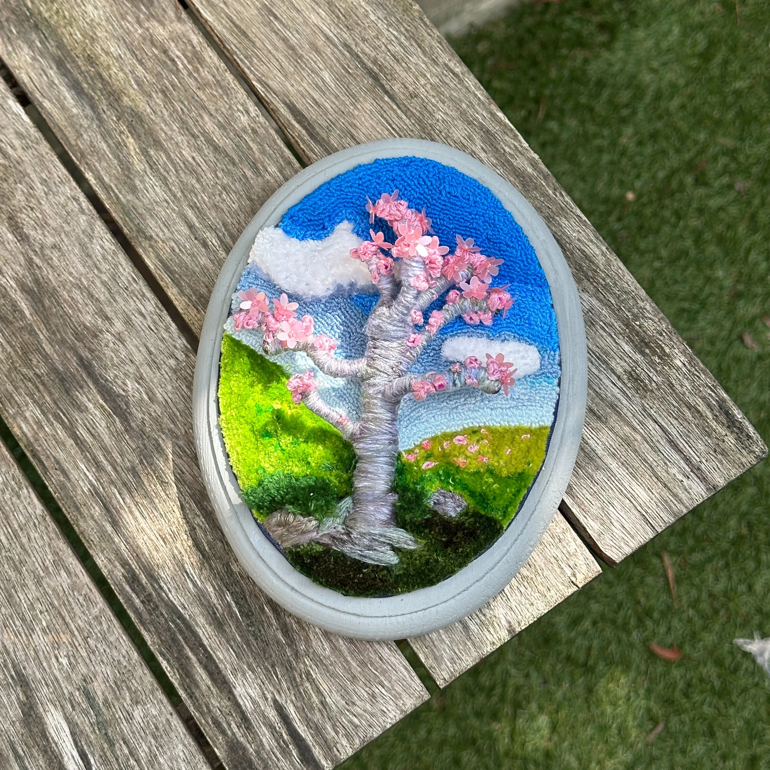 Decorative oval art piece featuring a cherry blossom tree with pink flowers, green grassy hills, blue sky, and white clouds, displayed on a wooden surface outdoors.