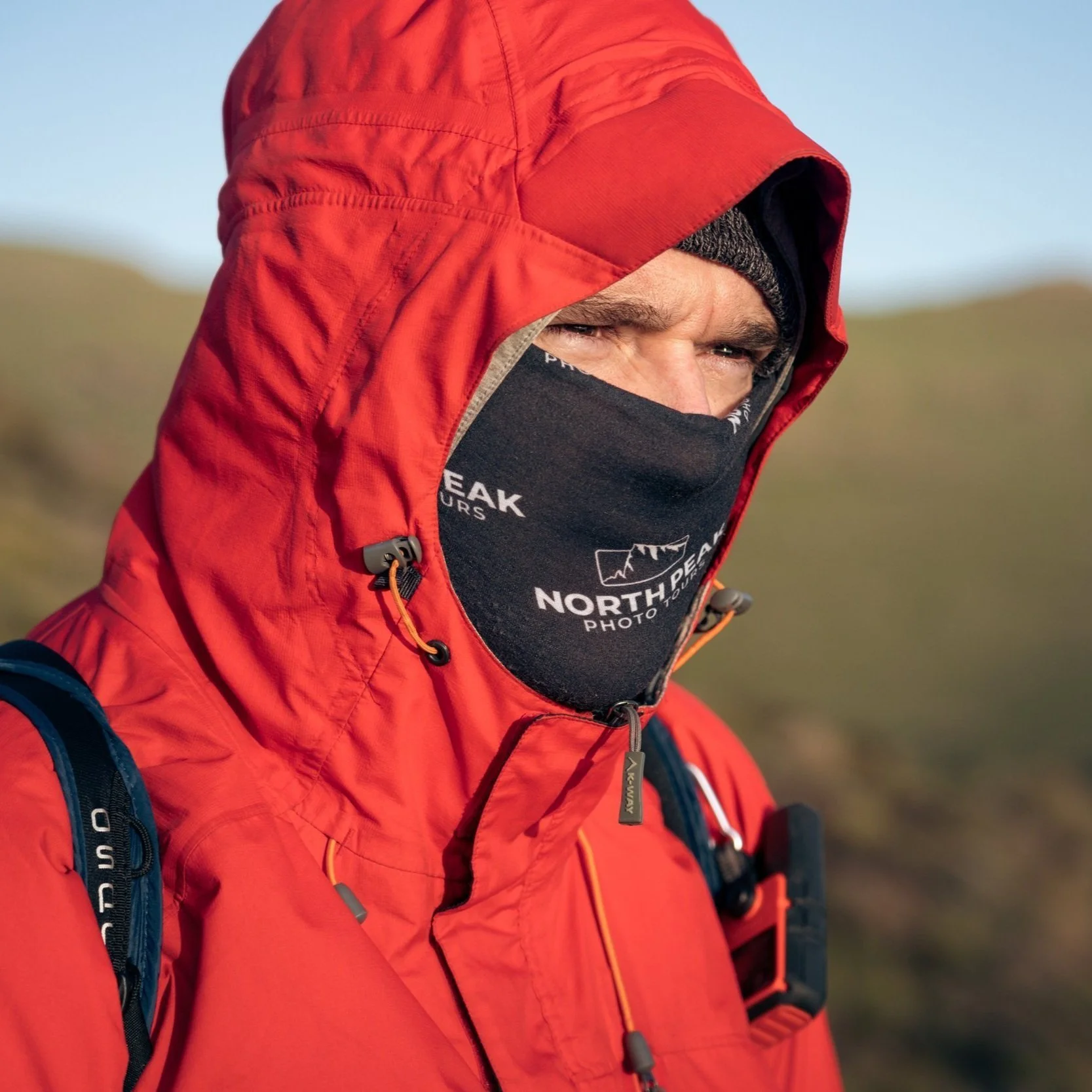 A man wearing a red jacket with the hood up and a black neck gaiter covering his face, outdoors on a cloudy day.