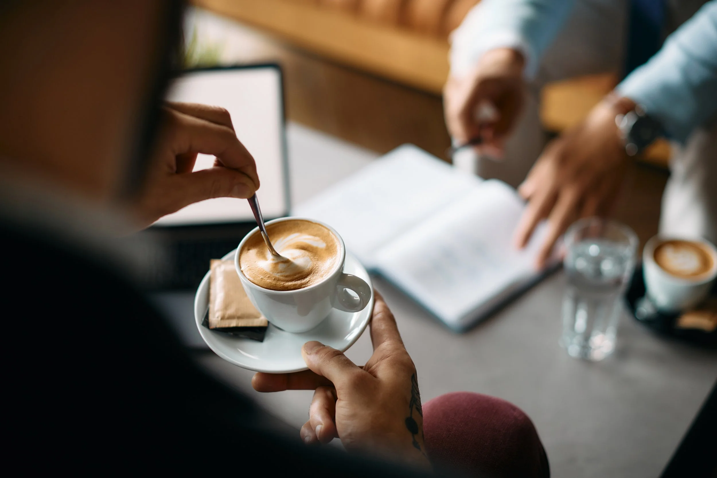 Person holding a tray with a cup of coffee, stirring it with a spoon, in a coffee shop or office setting.