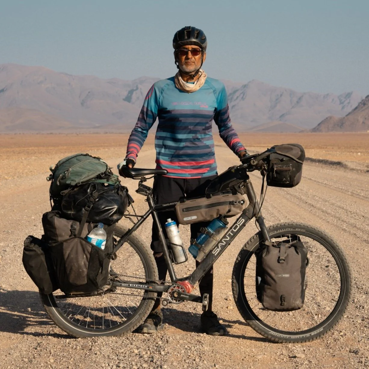 A man posing with a loaded mountain bike on a dirt trail in a desert with mountain ranges in the background, wearing a helmet, sunglasses, and athletic clothing.