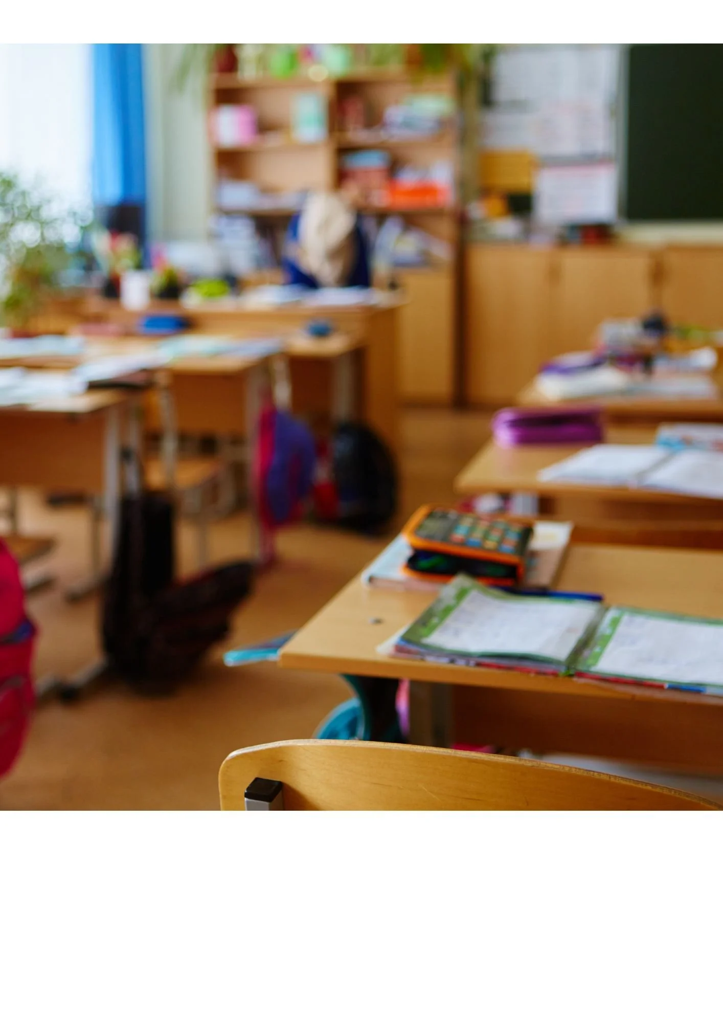 A classroom with wooden desks, open notebooks, and school supplies on the desks. Blurred background of shelves with books and a teacher at the blackboard.