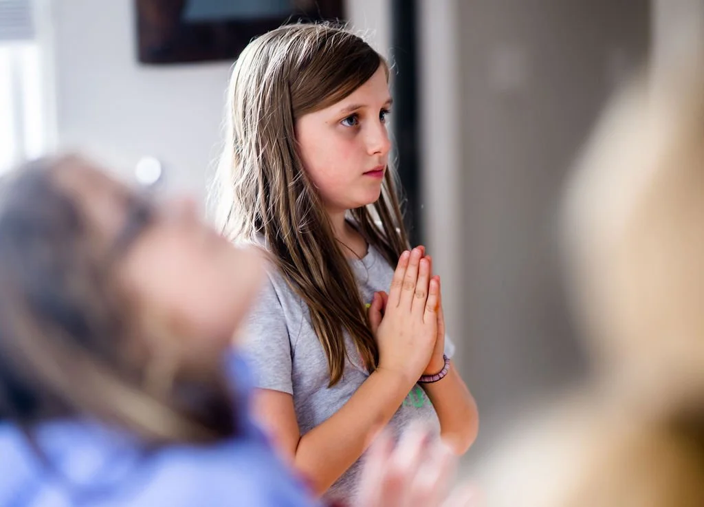 A young girl with long brown hair and blue eyes, wearing a light gray shirt, holds her hands together in a praying position in front of her chest, standing indoors.