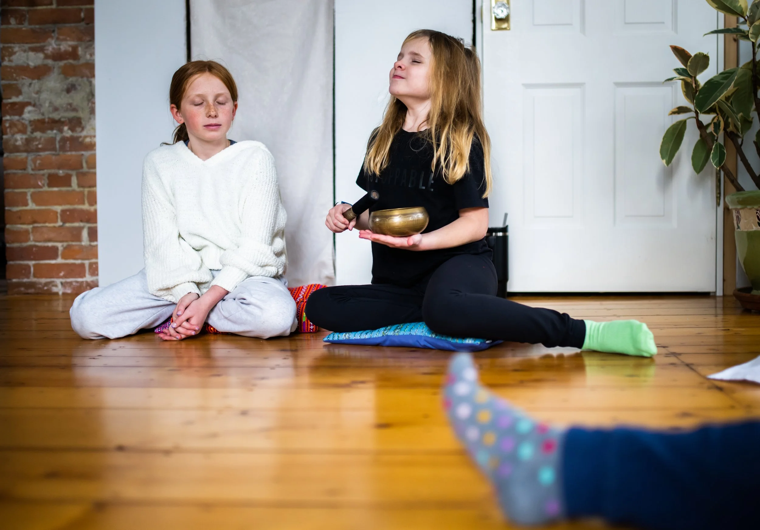 Two young girls sitting cross-legged on a wooden floor. One girl has red hair, wearing a white sweater and gray pants, with eyes closed. The other girl, with blond hair, is holding a singing bowl and a mallet, eyes closed, kneeling on a cushion. A third child's foot, wearing a colorful sock, is visible in the foreground.