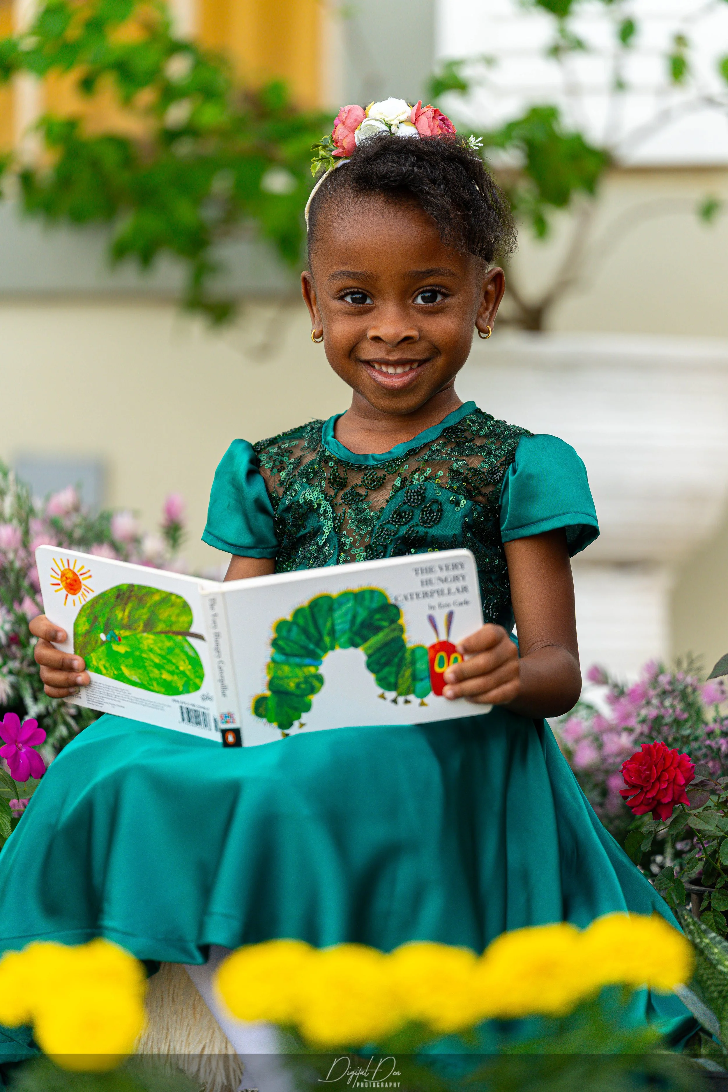 A young girl with a pink headband smiling while sitting on a green park bench and holding a wooden toy camera, outdoors with blurred flowers and greenery in the background.