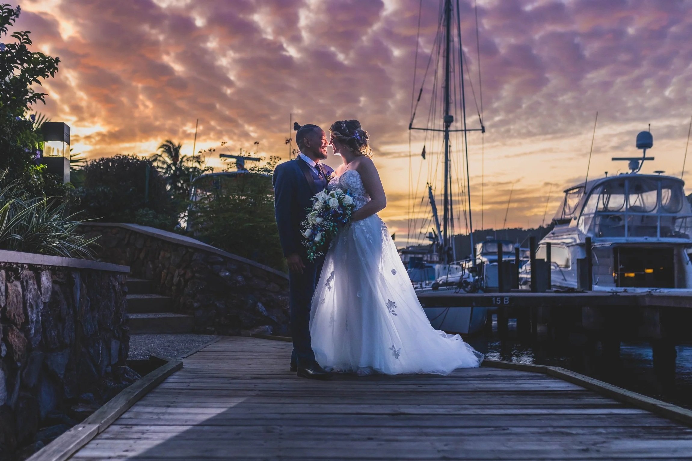 Bride and groom standing on a wooden dock by boats during sunset.