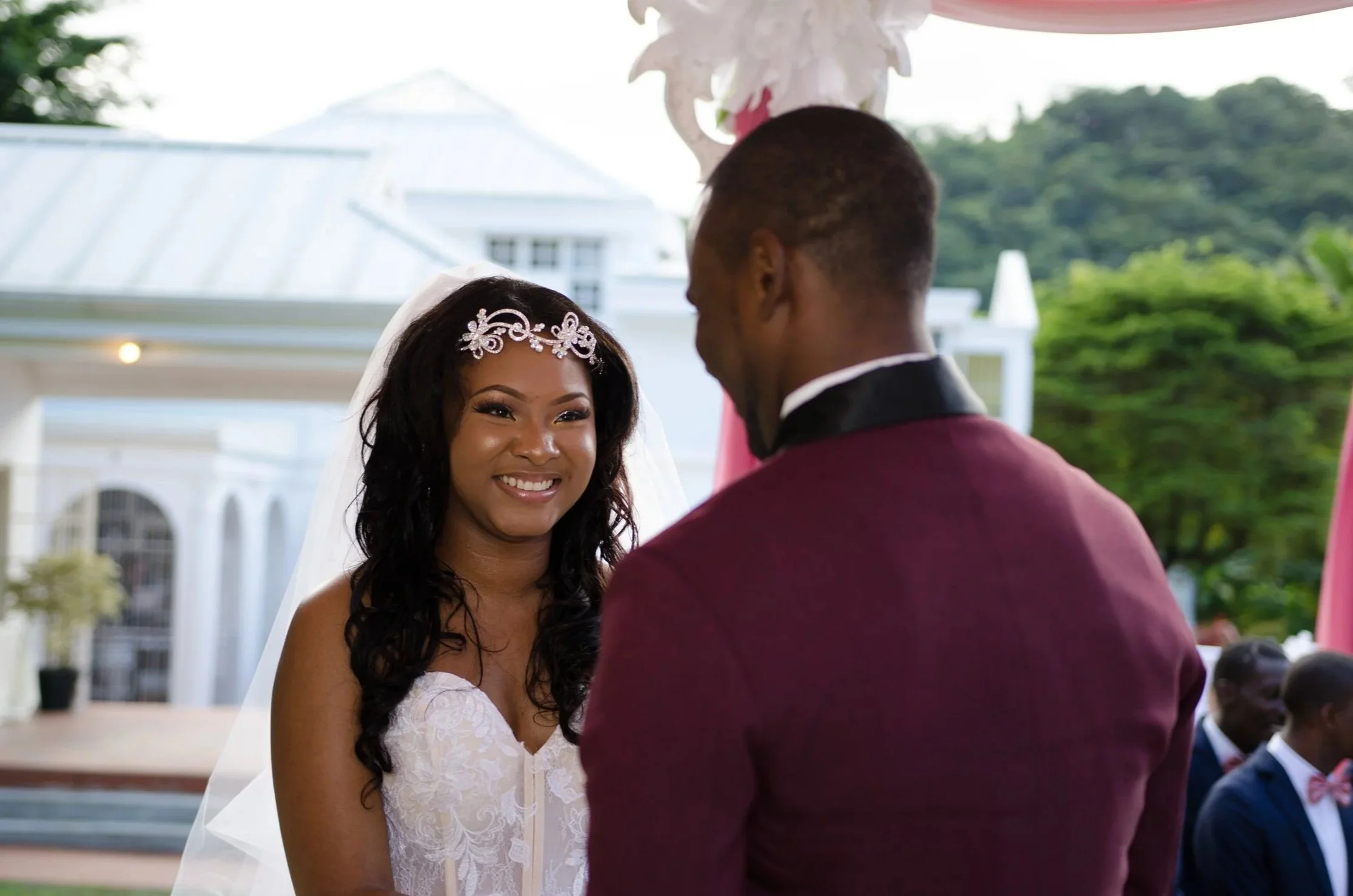 A bride and groom standing outside during their wedding ceremony, with the bride smiling at the groom, who is facing her.