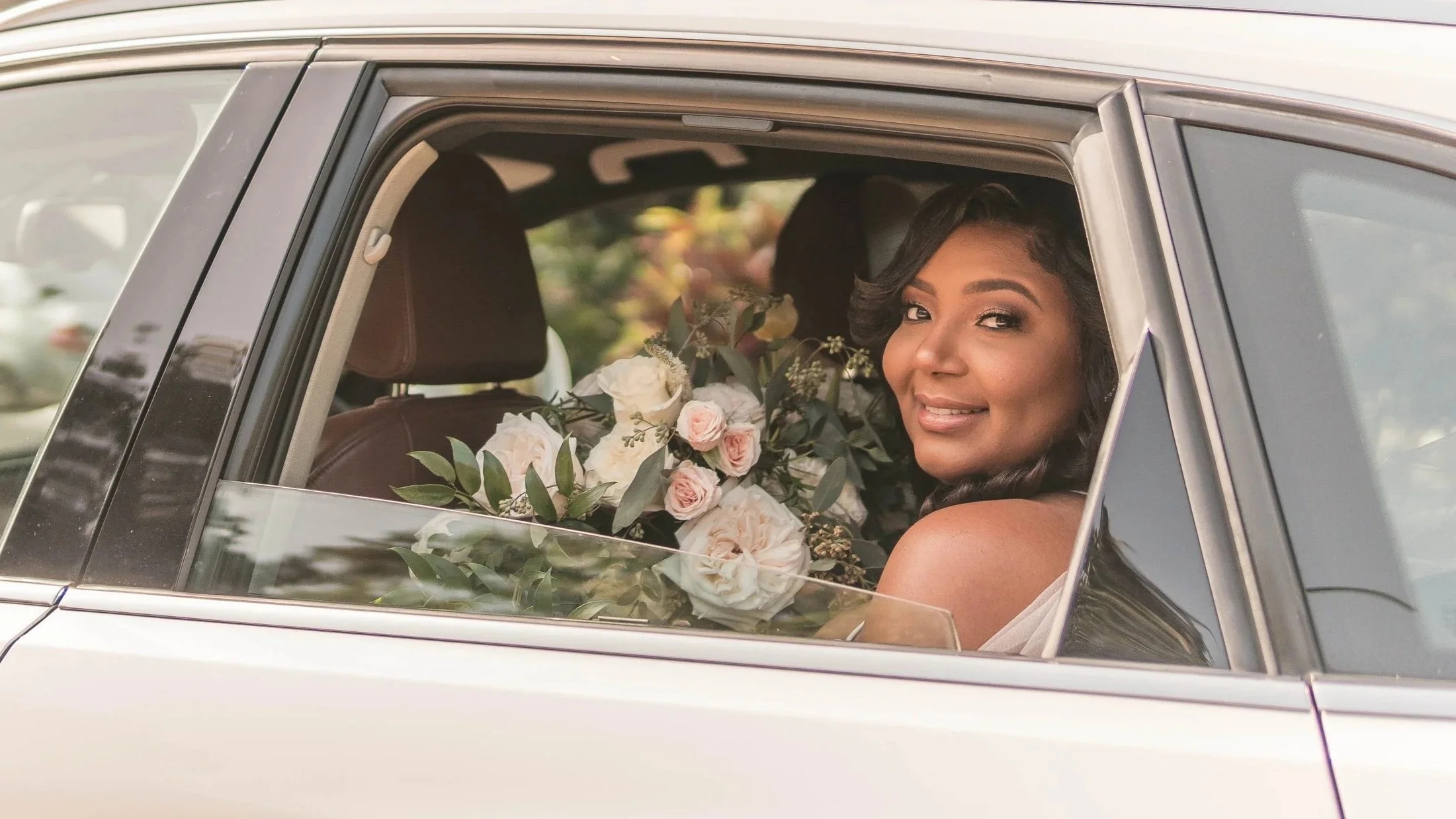 A woman sitting in a car holding a bouquet of white and pink roses, smiling and looking out the window.