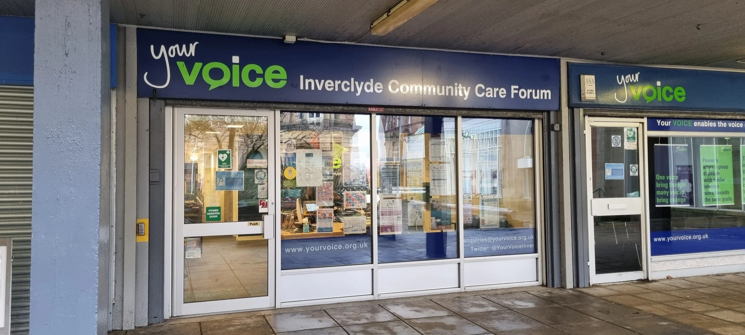Front view of Your Voice Inverclyde Community Care Forum building with glass doors and posters. The blue sign displays "Your Voice Inverclyde Community Care Forum."