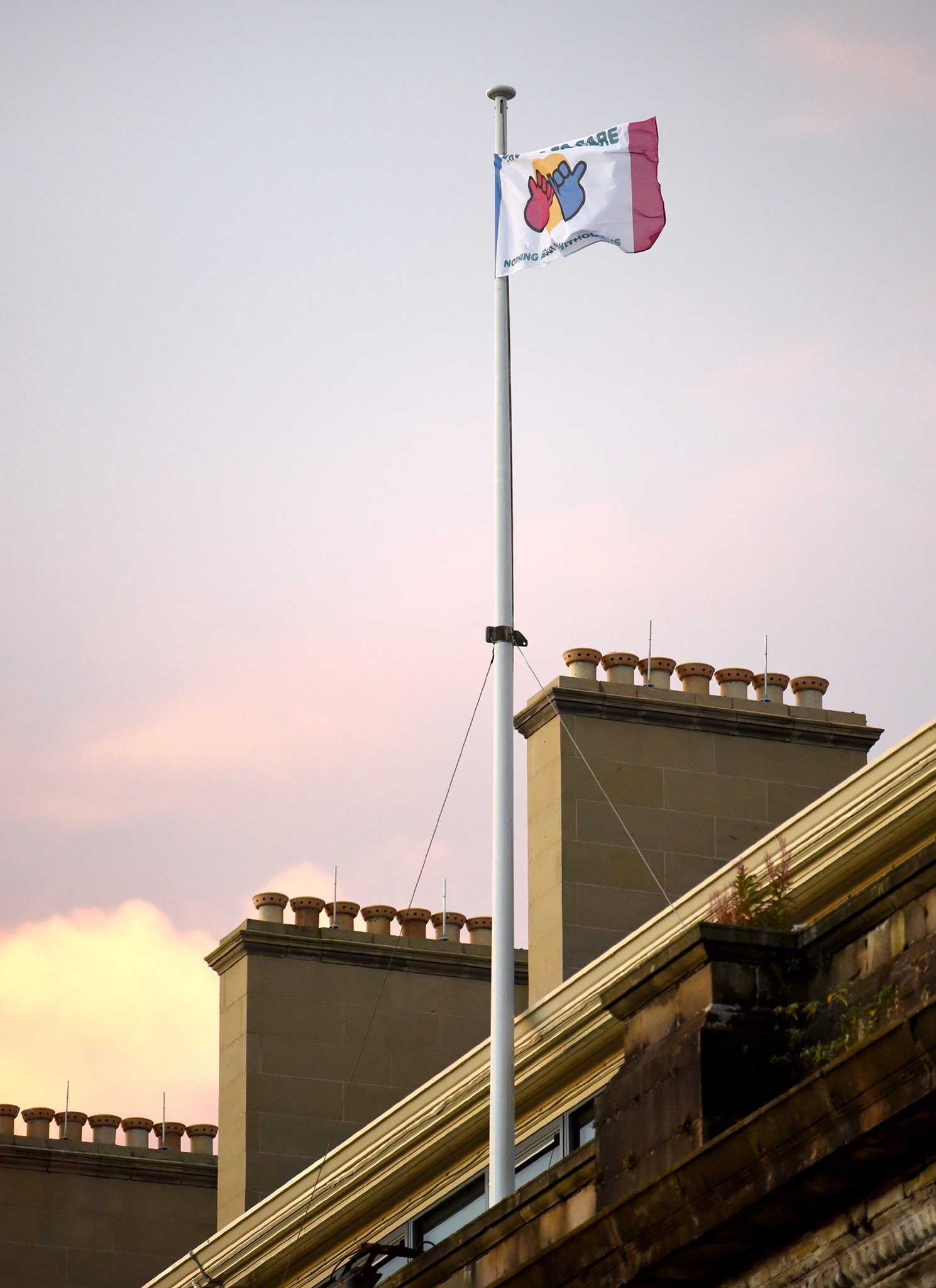 A flag with a colorful design on a pole, fluttering in the wind, against a background of a building with brick chimney stacks and a cloudy sky at sunset.