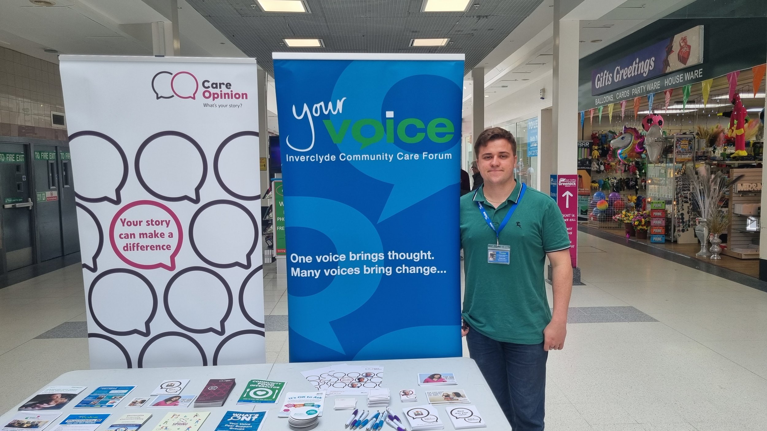 A person standing next to exhibition displays for Care Opinion and Your Voice Inverclyde Community Care Forum in a mall. The table in front has brochures and pens. The background shows a store named Gifts Greetings.