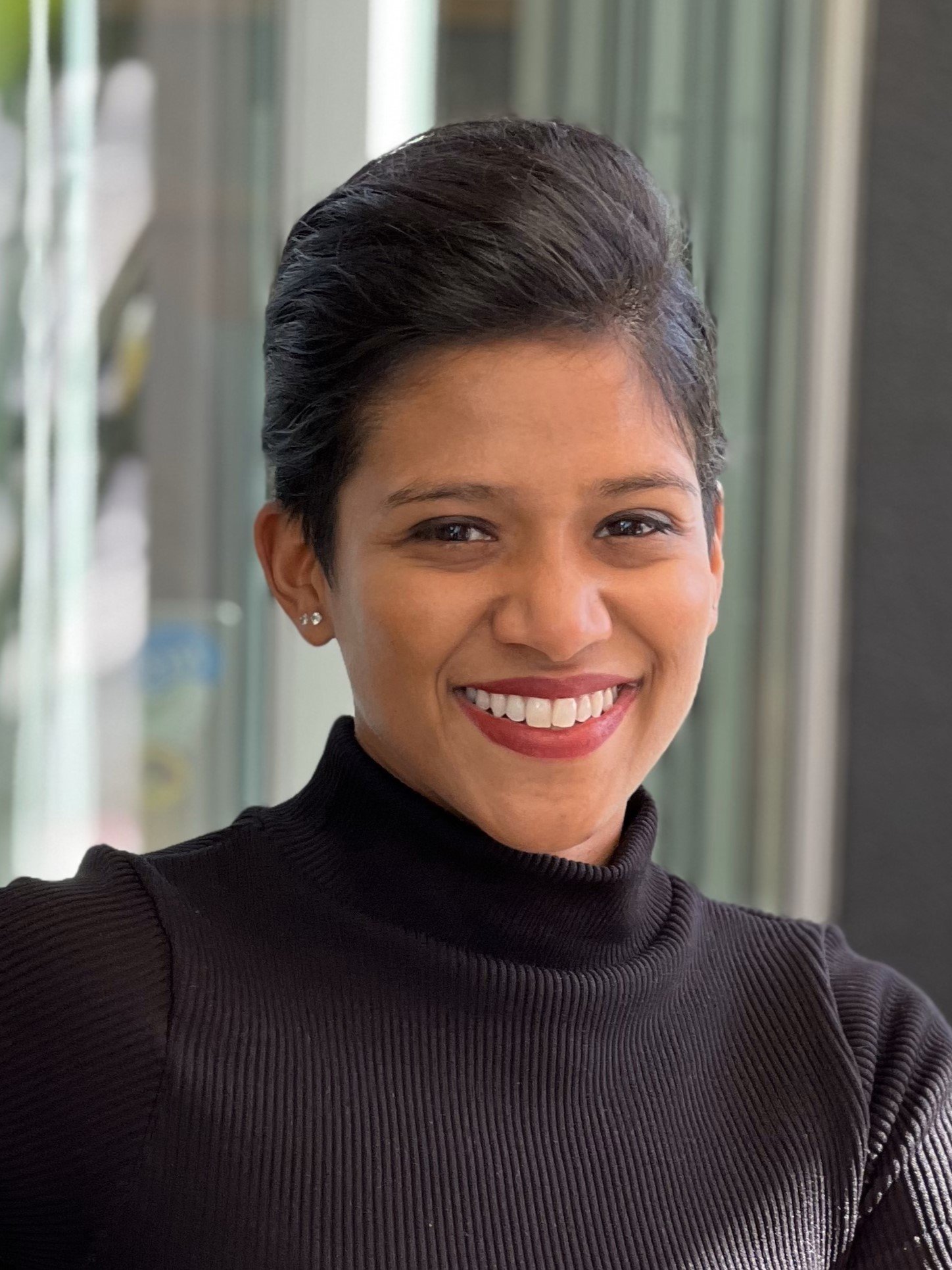 A smiling woman with short black hair wearing a black turtleneck sweater and small earrings, standing indoors near a window.