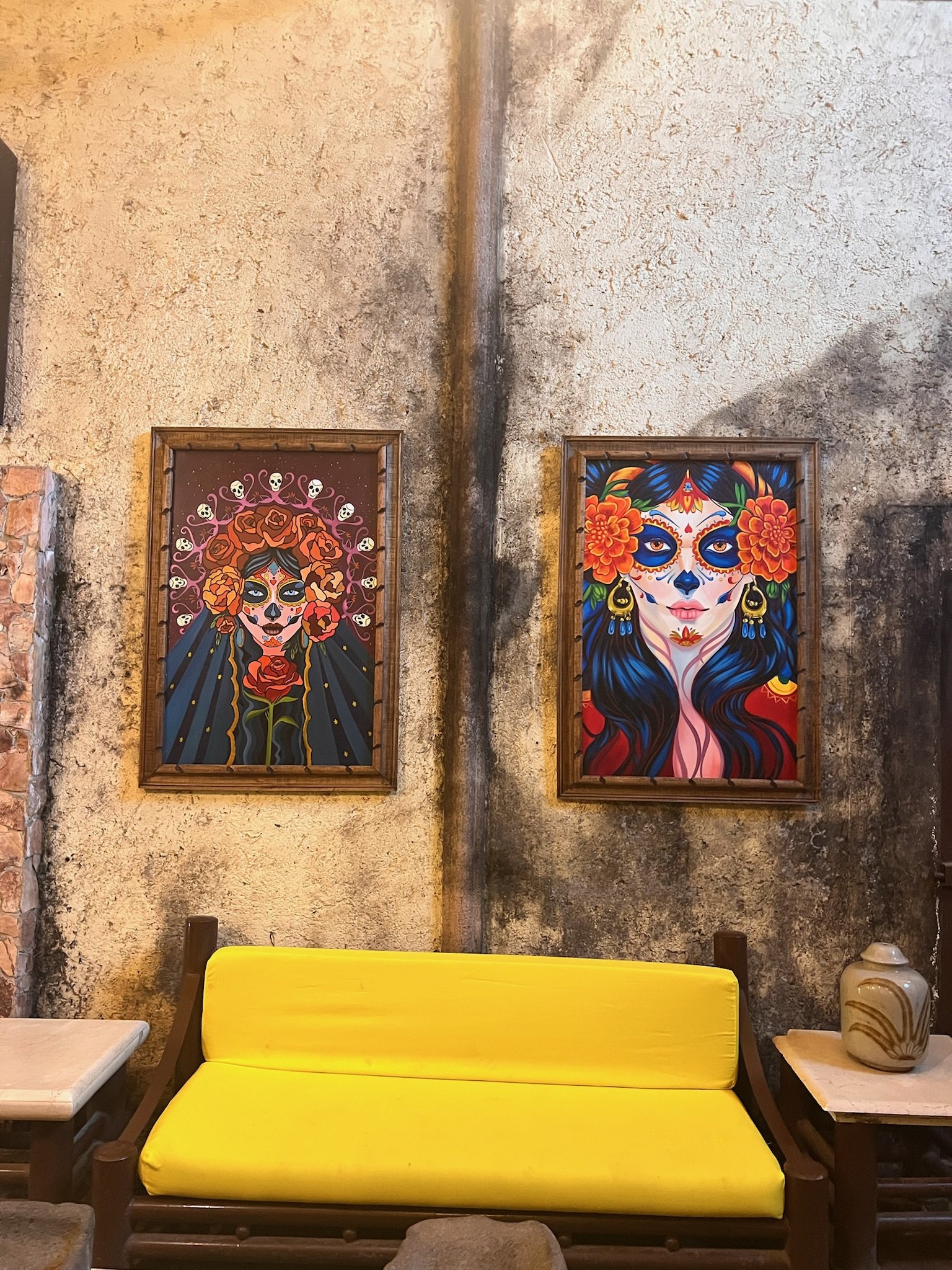 Wall with two framed Day of the Dead paintings above a yellow bench, rustic texture background.