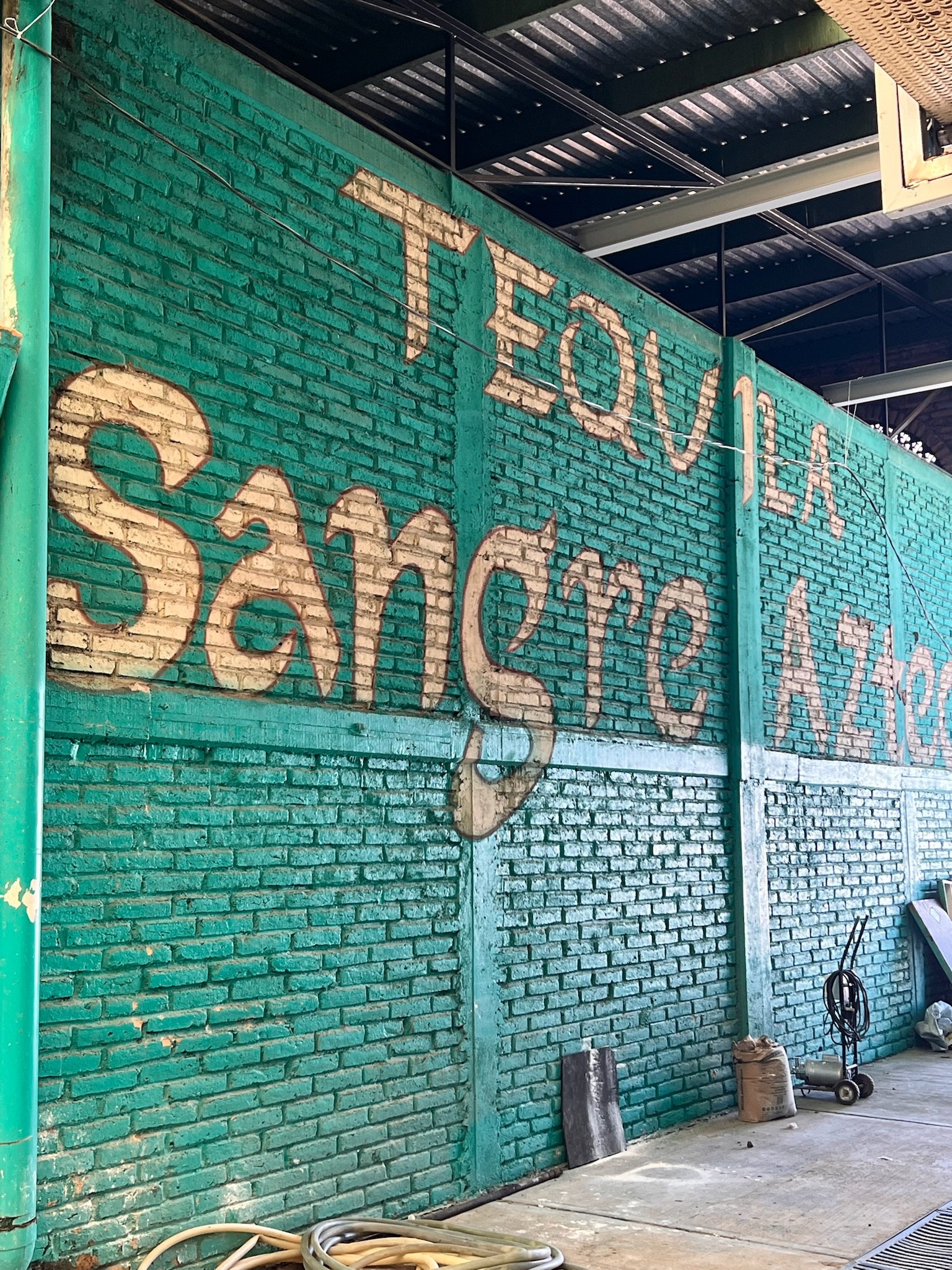 Teal brick wall with "Tequila Sangre Azteca" painted on it, inside an industrial building. Hoses and metal objects are on the floor.