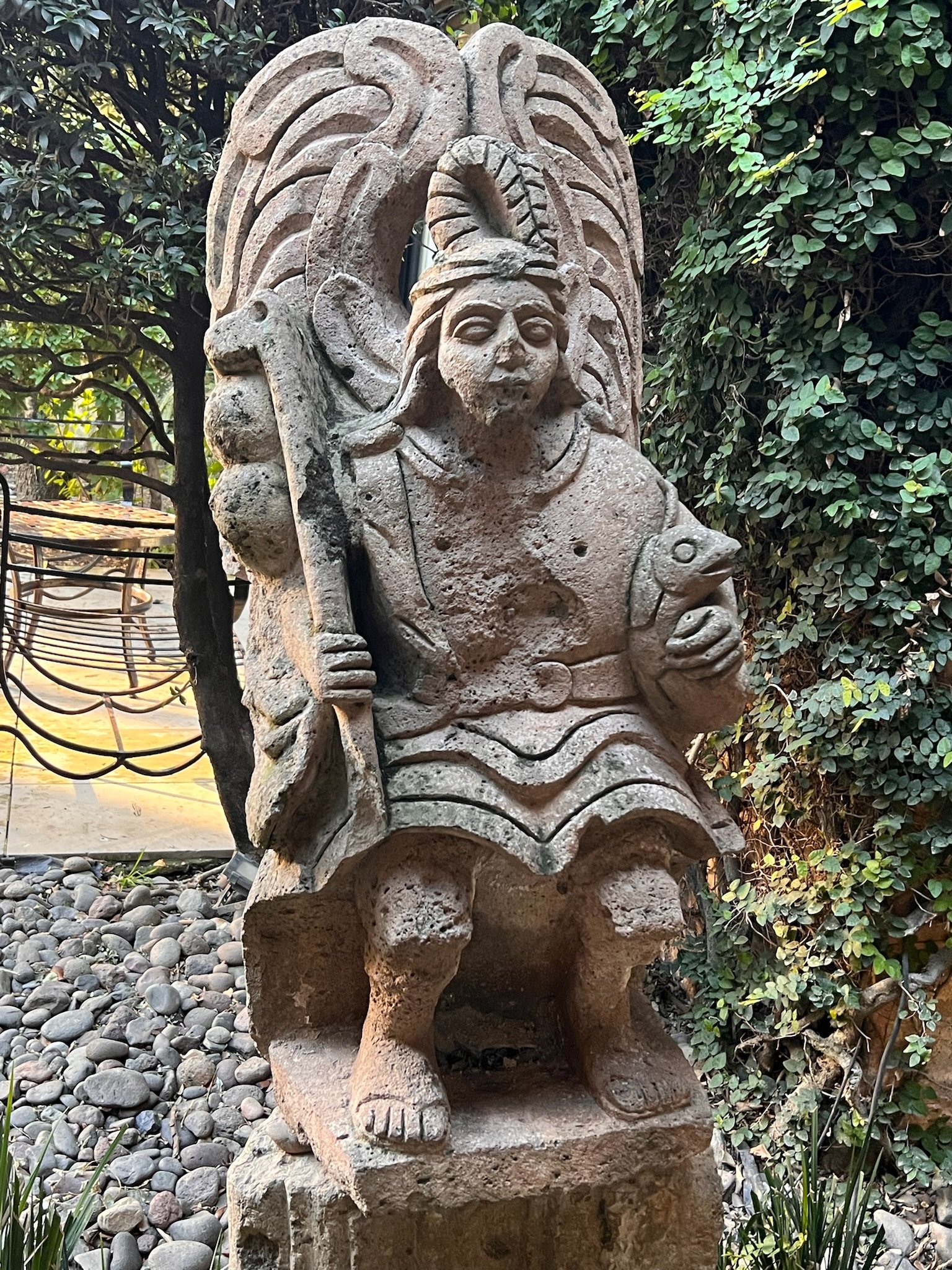 Stone statue of an Aztec or Maya warrior with headdress and shield, surrounded by foliage in a garden setting.