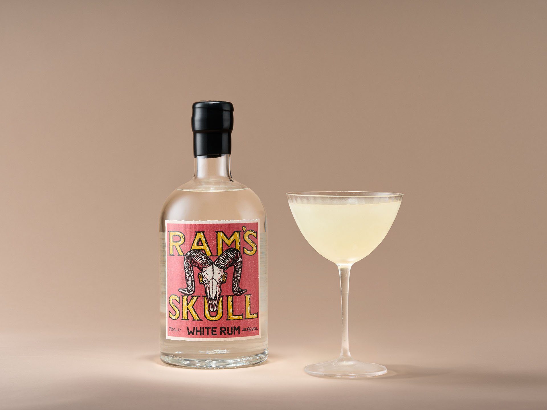 A bottle of Ram's Skull White Rum next to a cocktail glass filled with a light-colored drink against a neutral background.